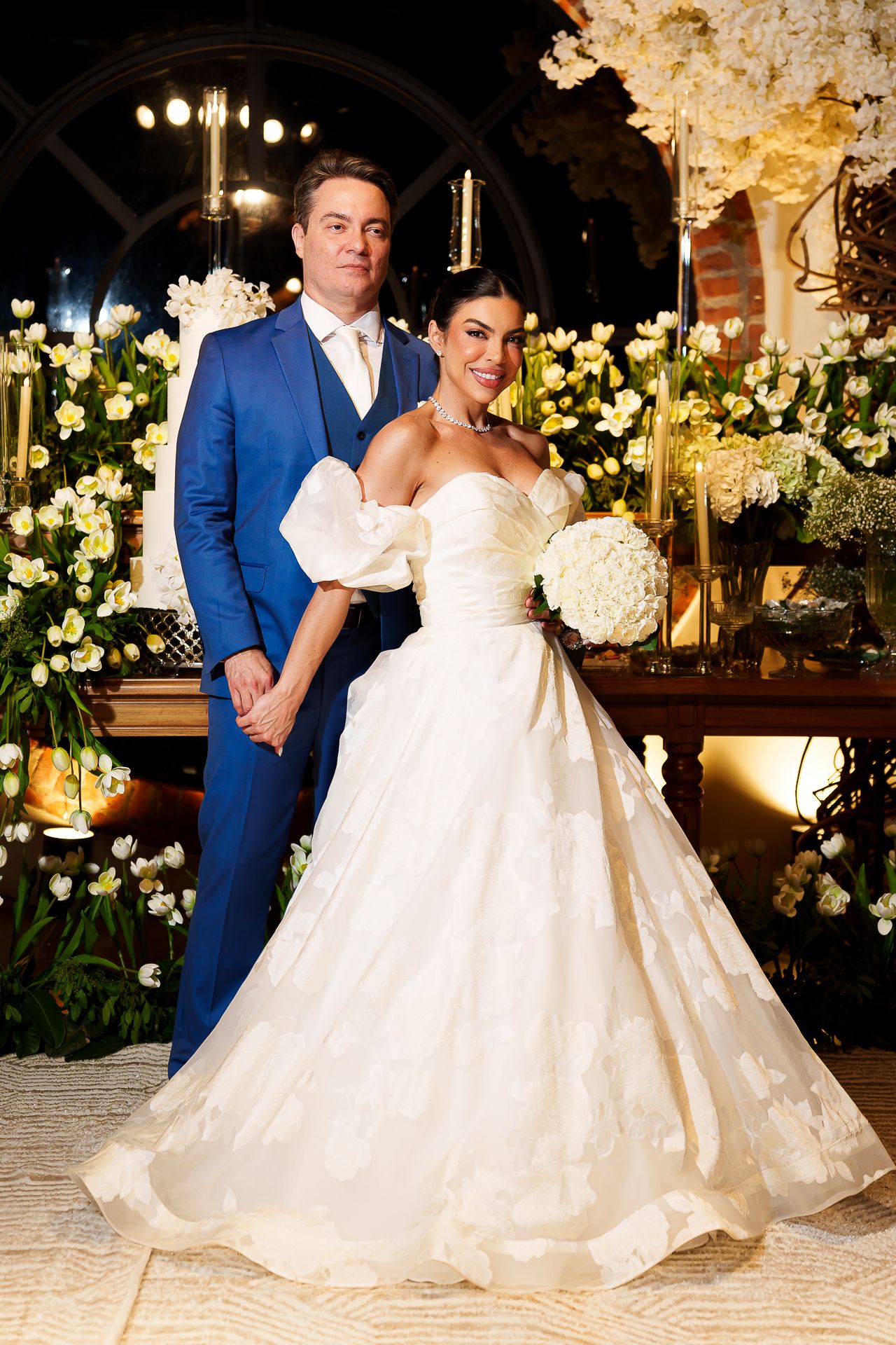 Bride in strapless ballgown holding white bouquet stands with groom in blue suit at elegant floral wedding reception.