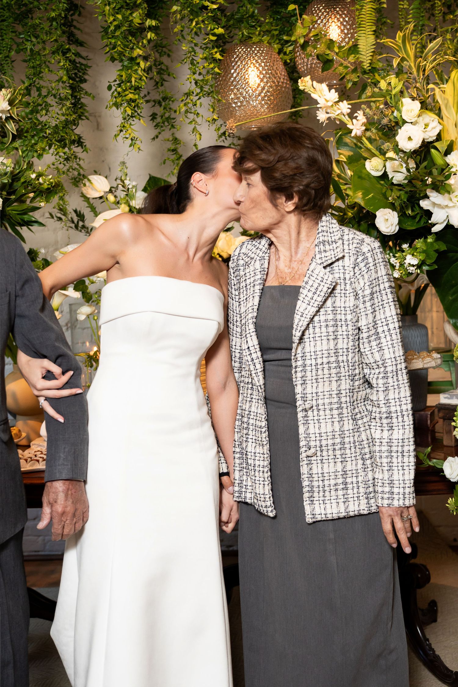 Bride in white strapless gown shares a kiss with an older woman in a tweed jacket at a wedding reception.
