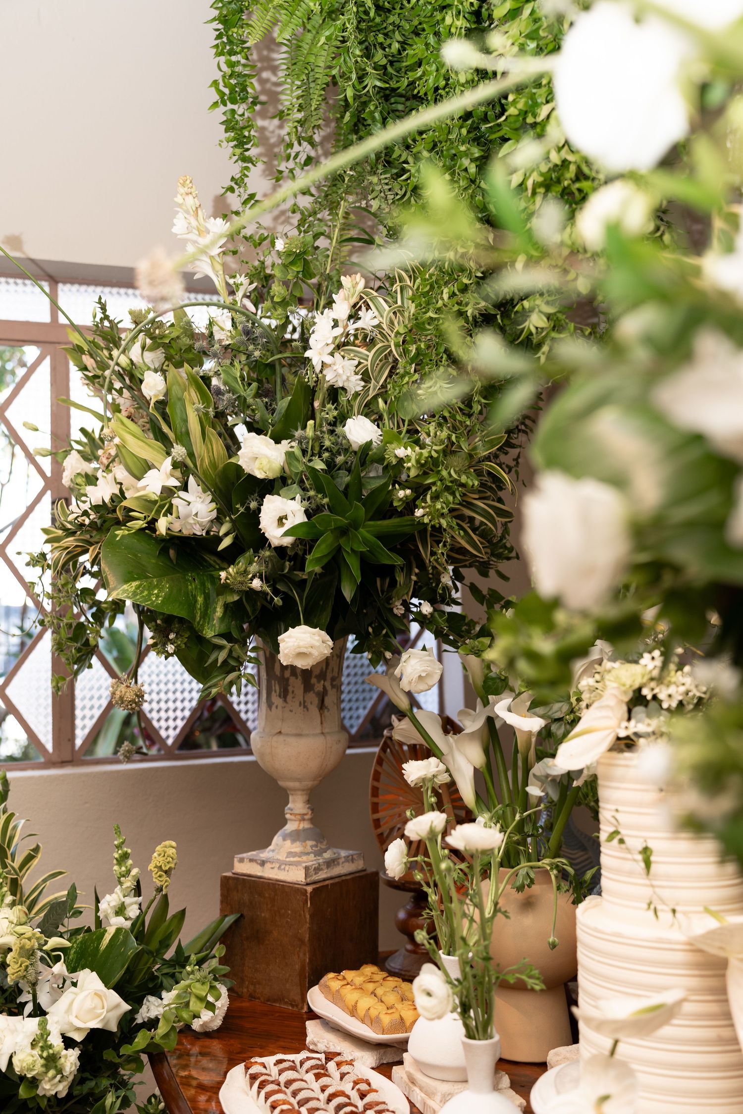 Elegant wedding dessert table with white floral arrangements, greenery, and a tiered cake in a garden setting.