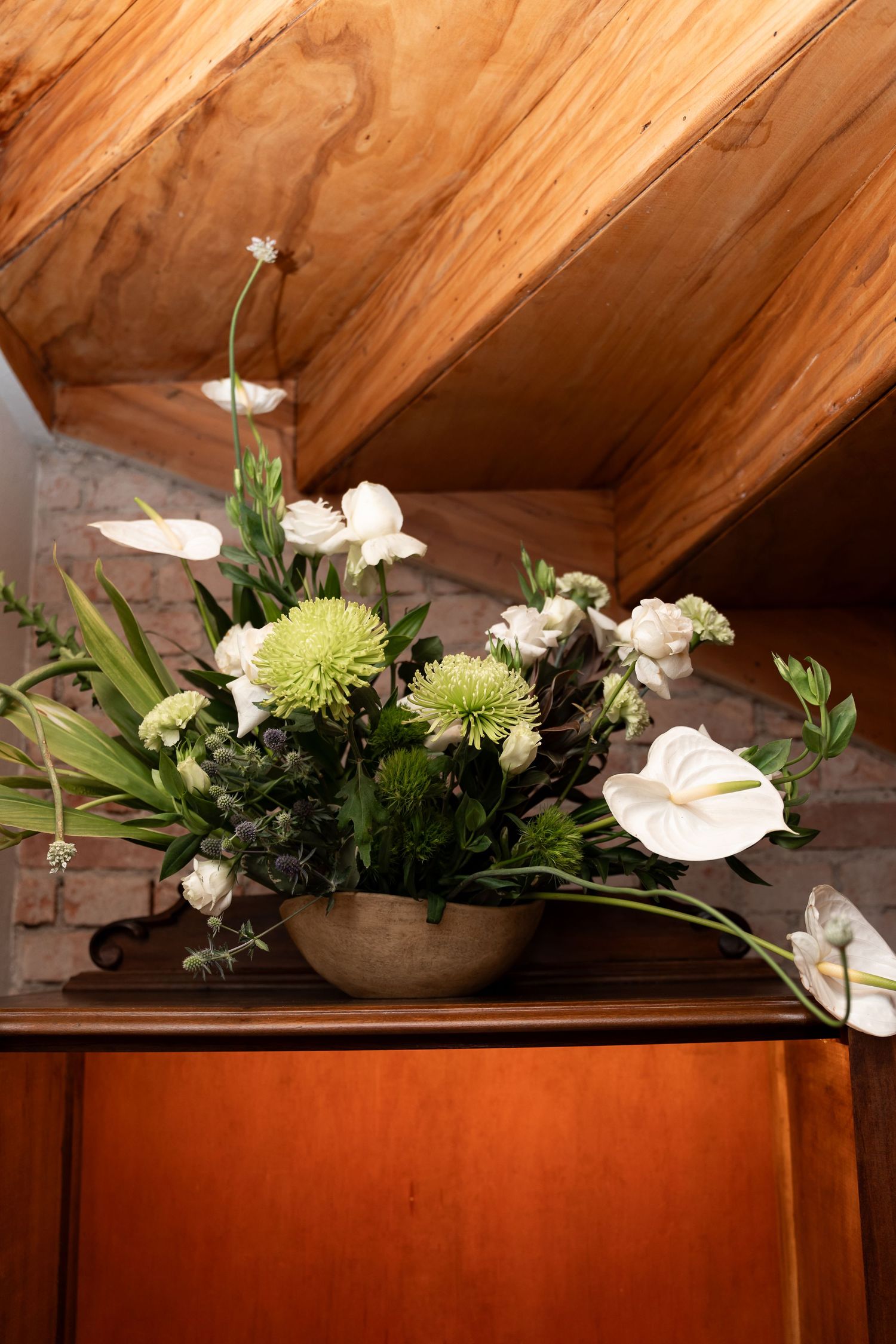 Elegant floral arrangement with white anthuriums, green chrysanthemums in a bowl vase on wooden cabinet under sloped ceiling.