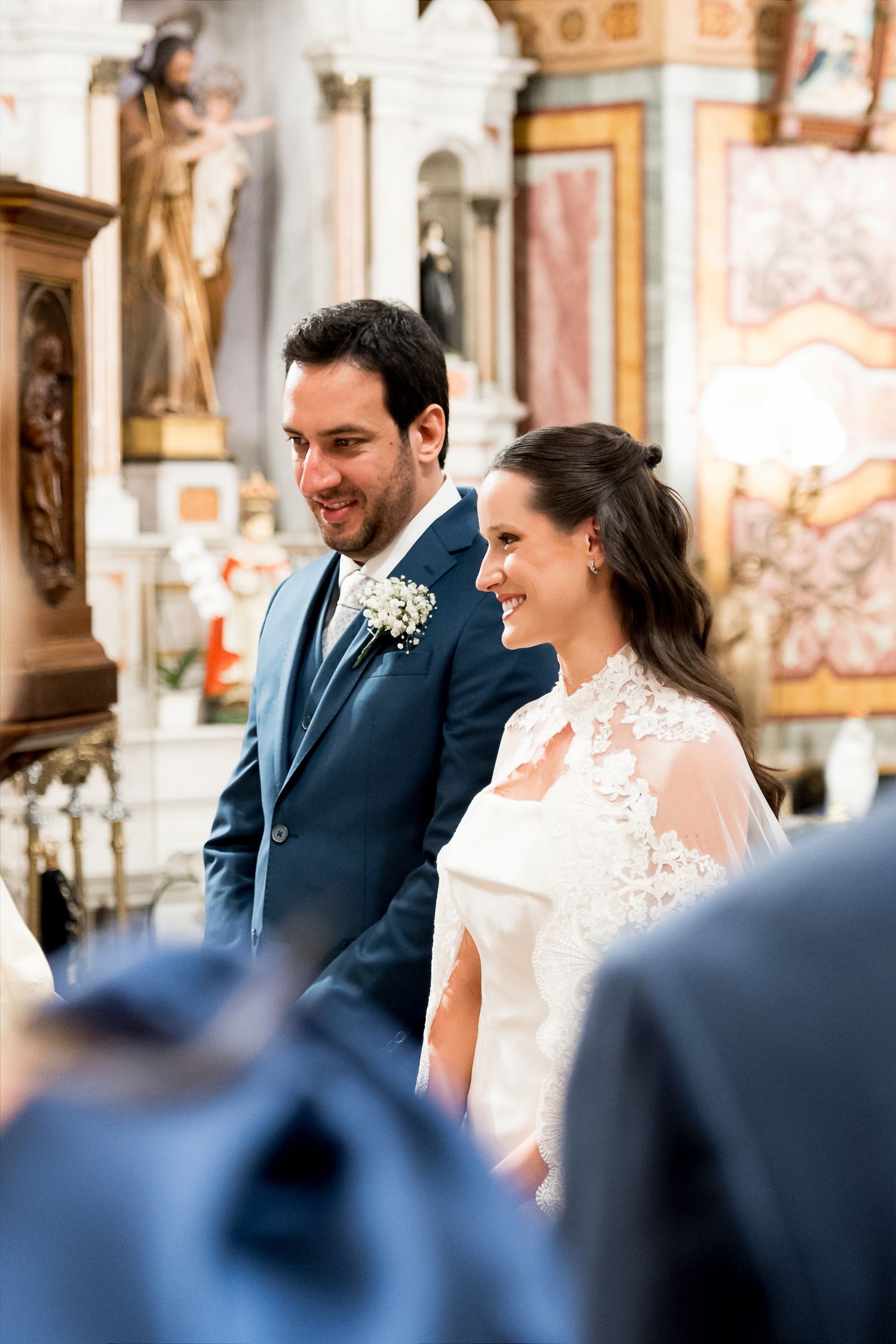 A smiling bride in lace wedding dress and groom in blue suit stand together during a church wedding ceremony.