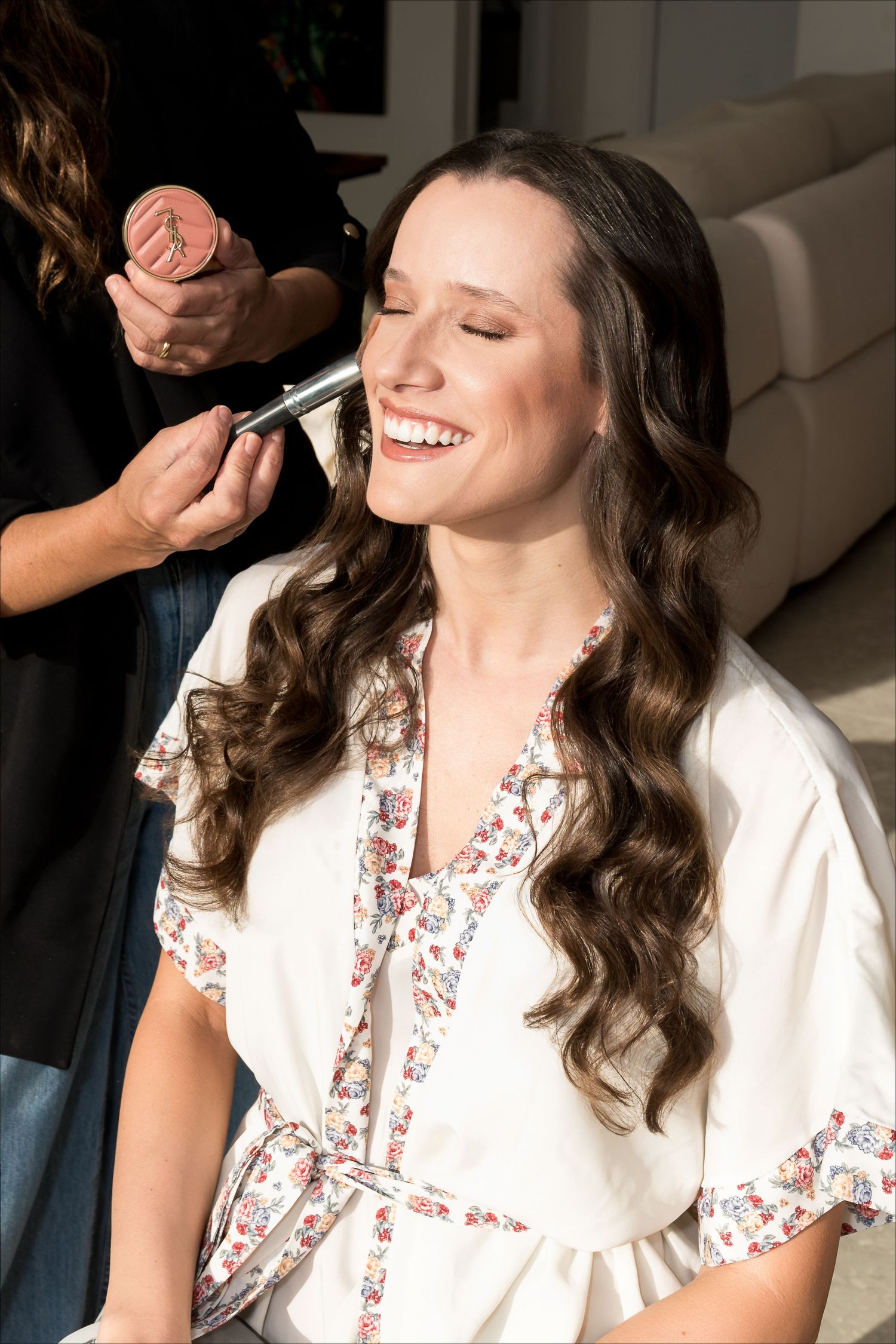 Smiling bride getting makeup applied by artist while wearing floral robe during wedding preparation.