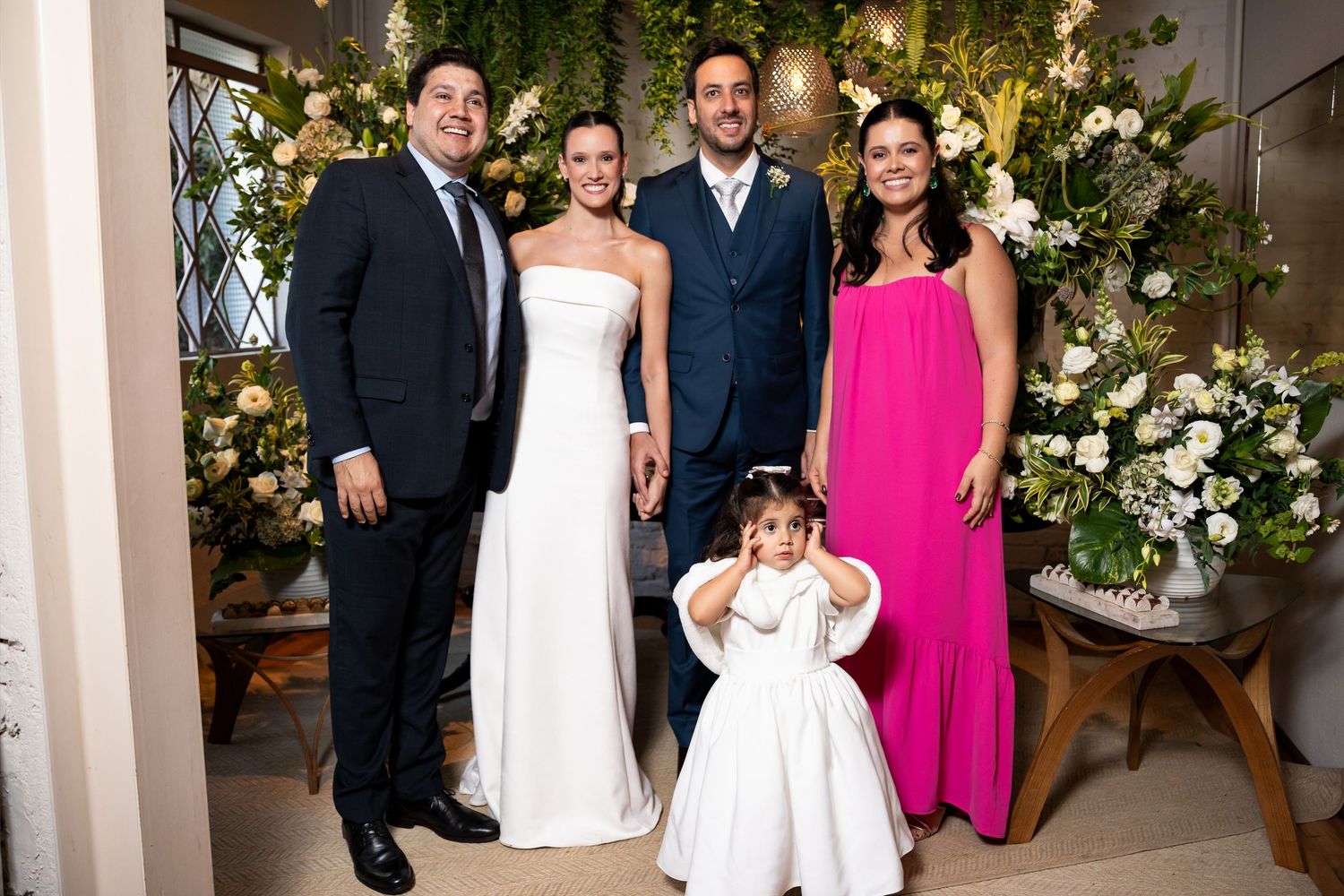 Wedding party poses with bride, groom, guests in pink dress, and flower girl before floral backdrop.