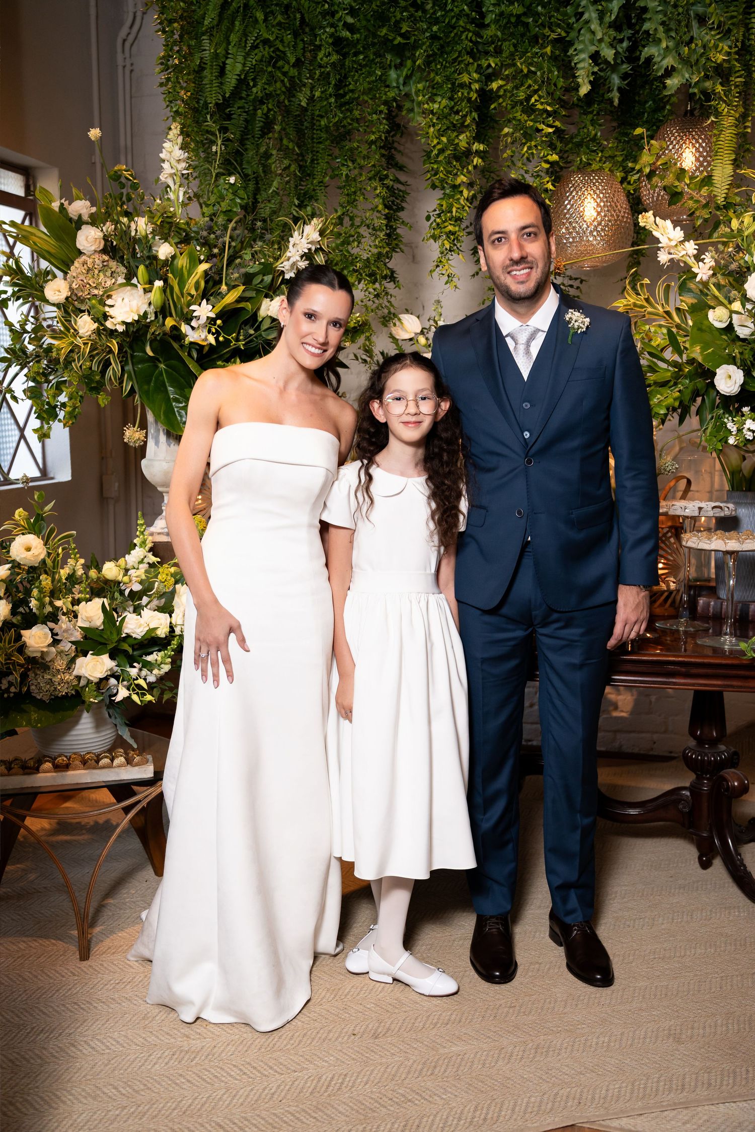 Bride and groom pose with young girl in white dress at elegant wedding reception with floral greenery backdrop.