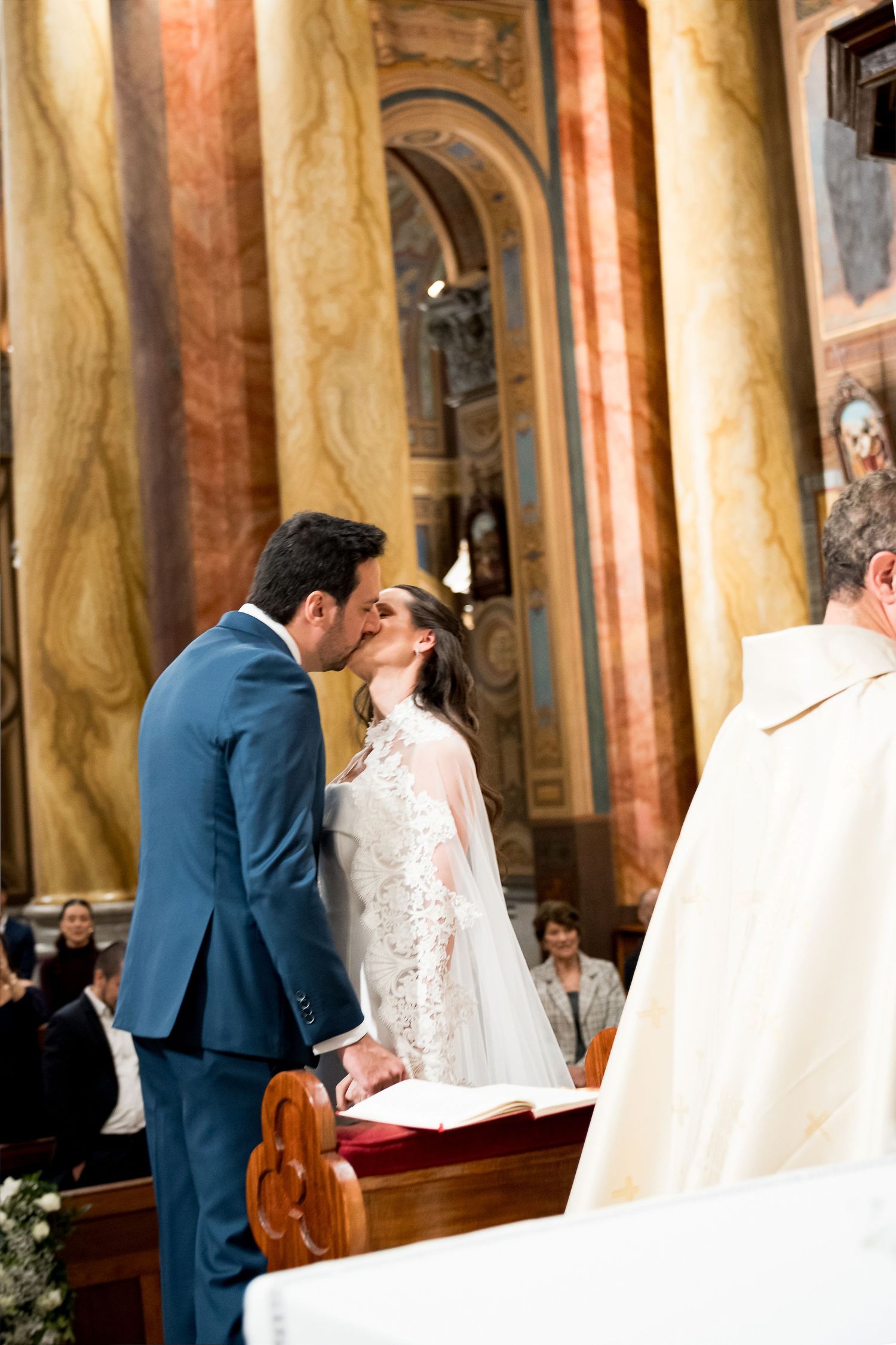 Bride and groom share first kiss at church altar during elegant wedding ceremony with marble columns.