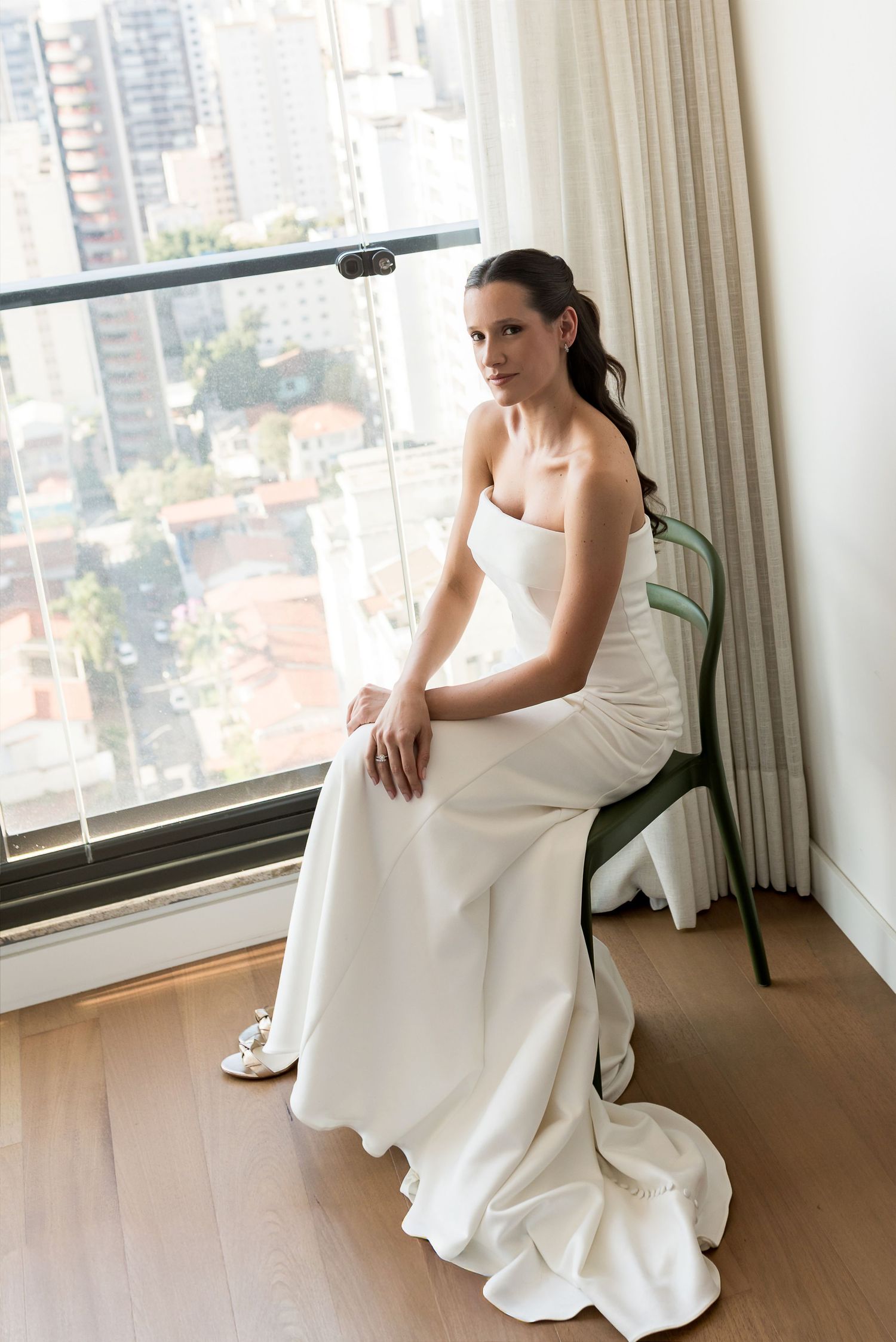 Bride in a strapless white gown sits gracefully on a green chair by a large window with city views.