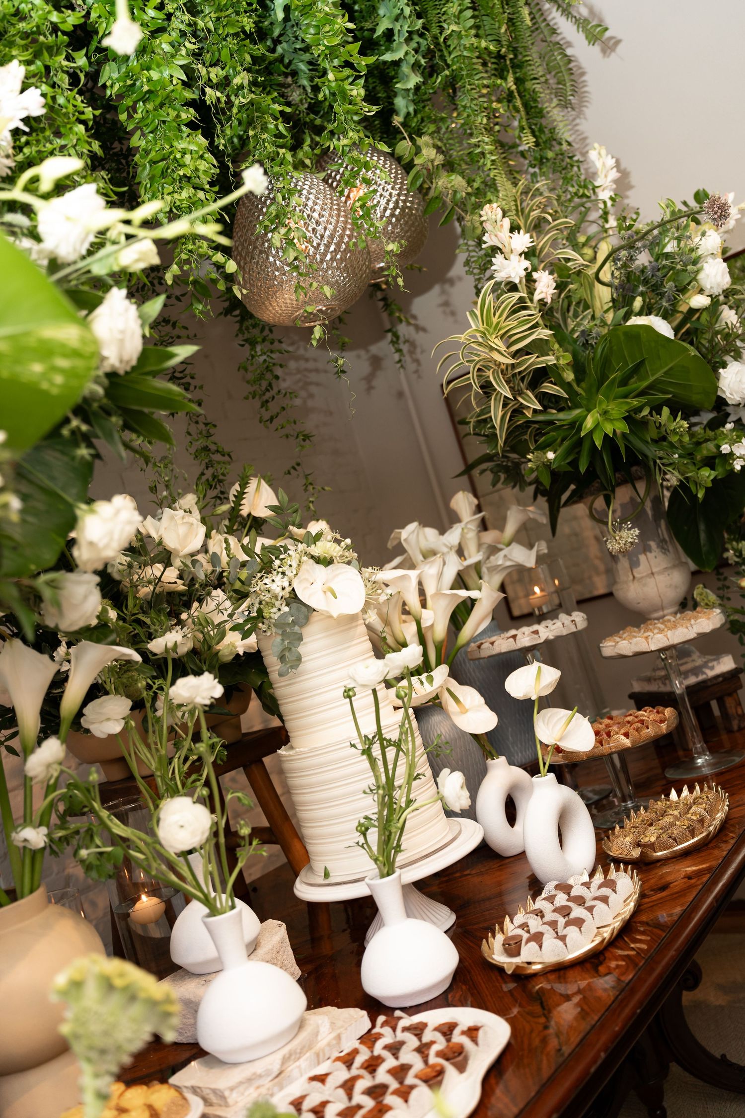 Elegant wedding dessert table with white cake, floral arrangements, and sweet treats surrounded by lush greenery.