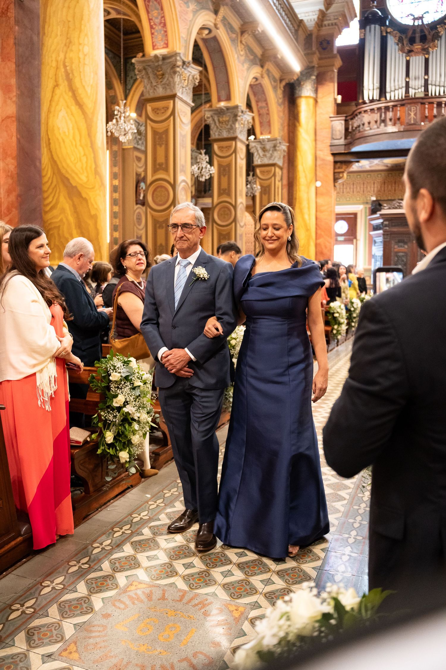 Couple walks down ornate church aisle in wedding ceremony, woman in navy gown, man in gray suit, guests standing.