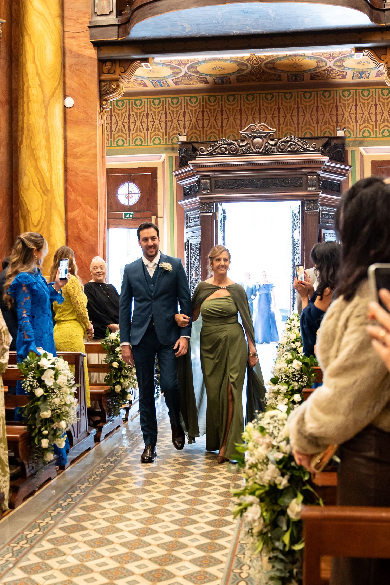Groom in navy suit walks bride in green gown down floral-lined church aisle as guests photograph the moment.