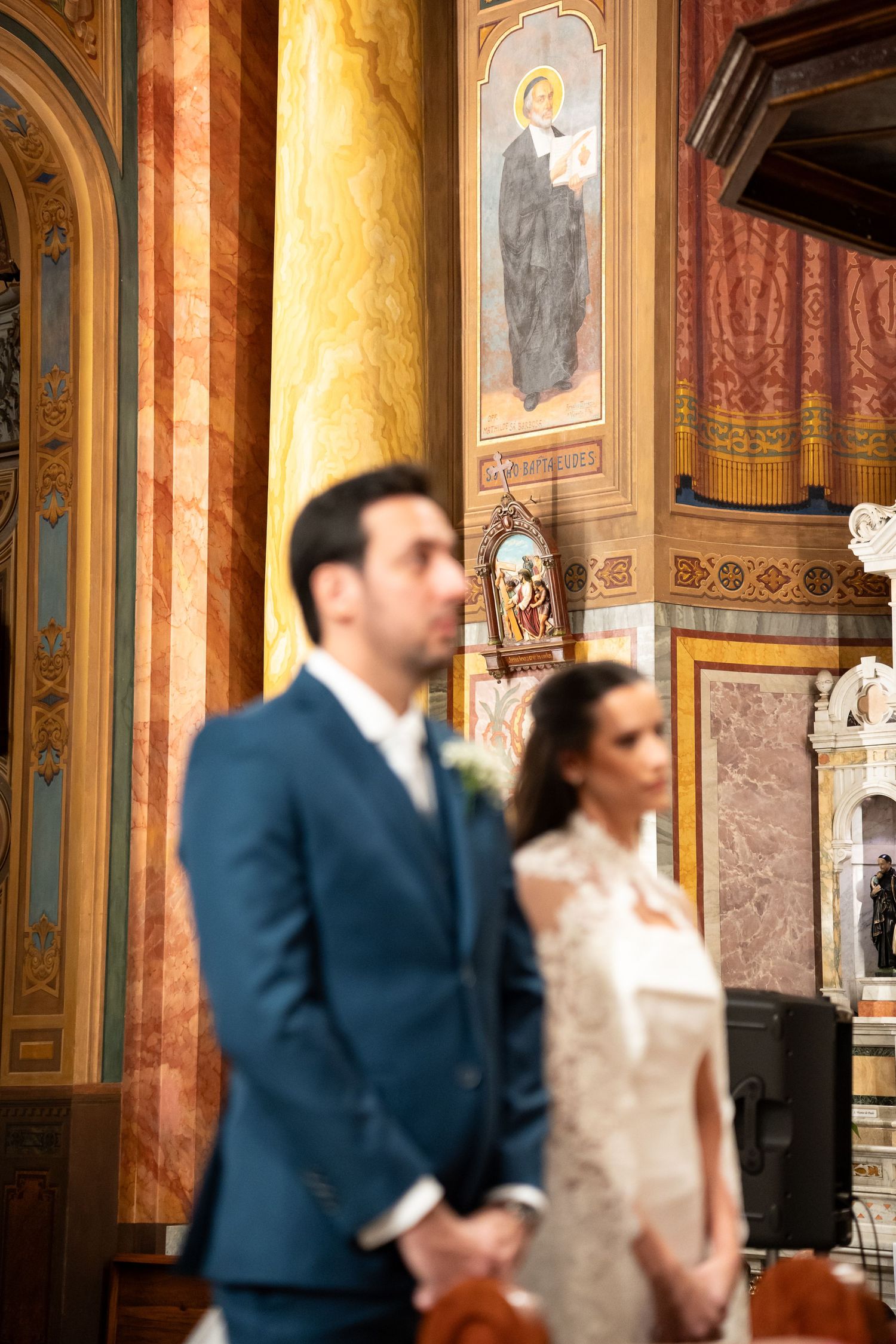 Bride and groom stand at the altar of an ornate church with gold columns and religious frescoes during wedding ceremony.