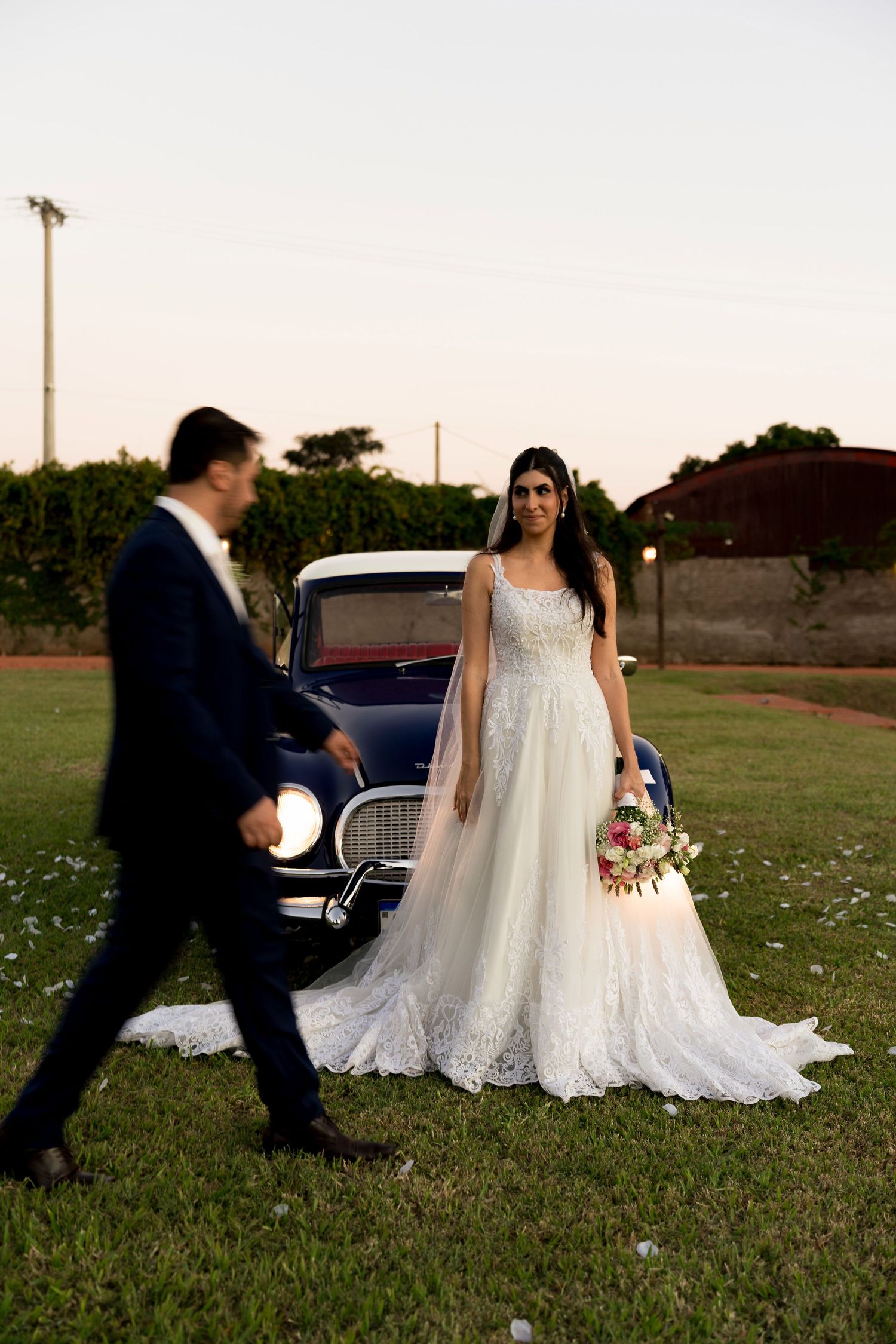 Bride in white lace gown holds bouquet by vintage car at dusk as groom in navy suit walks toward her.