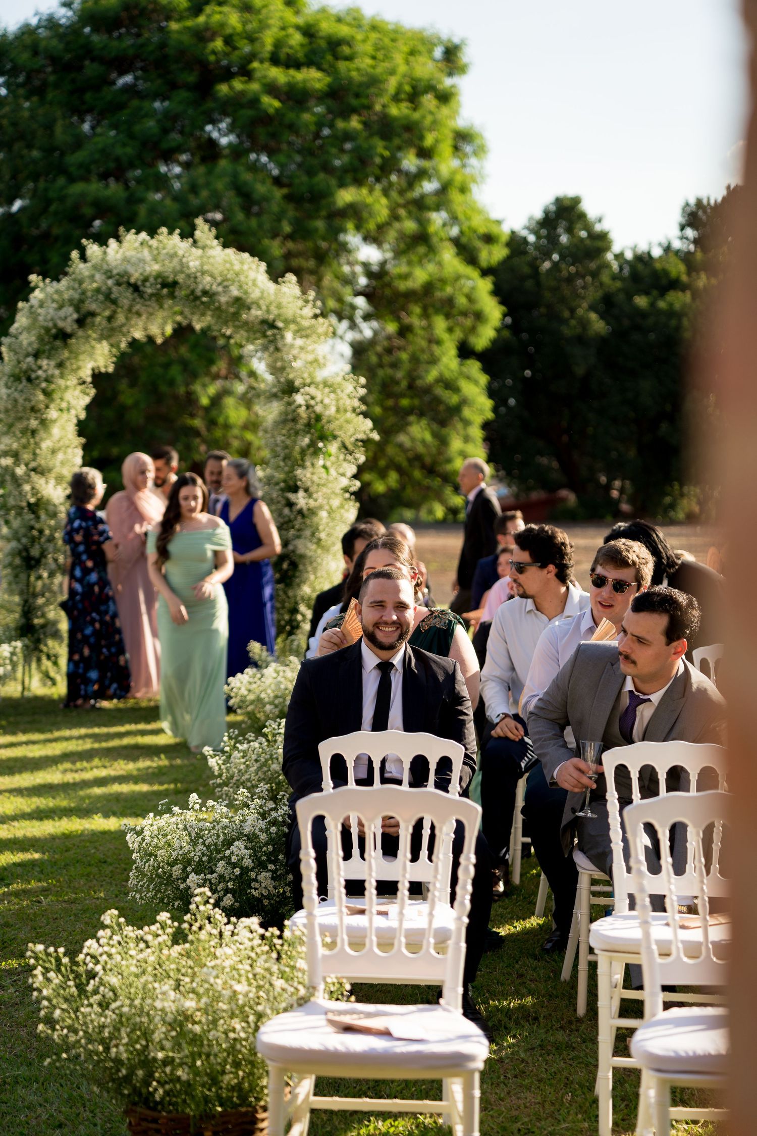 Outdoor wedding ceremony with guests seated on white chairs near a floral arch surrounded by lush greenery.