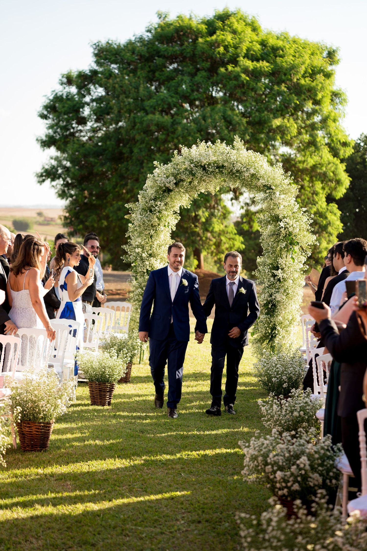 Two grooms walk hand-in-hand down a floral arch aisle at an outdoor wedding ceremony.