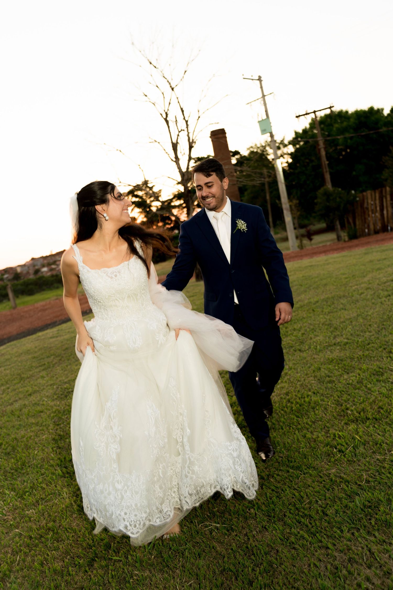 A joyful bride in a lace gown and groom in navy suit walk hand-in-hand on green grass at sunset.