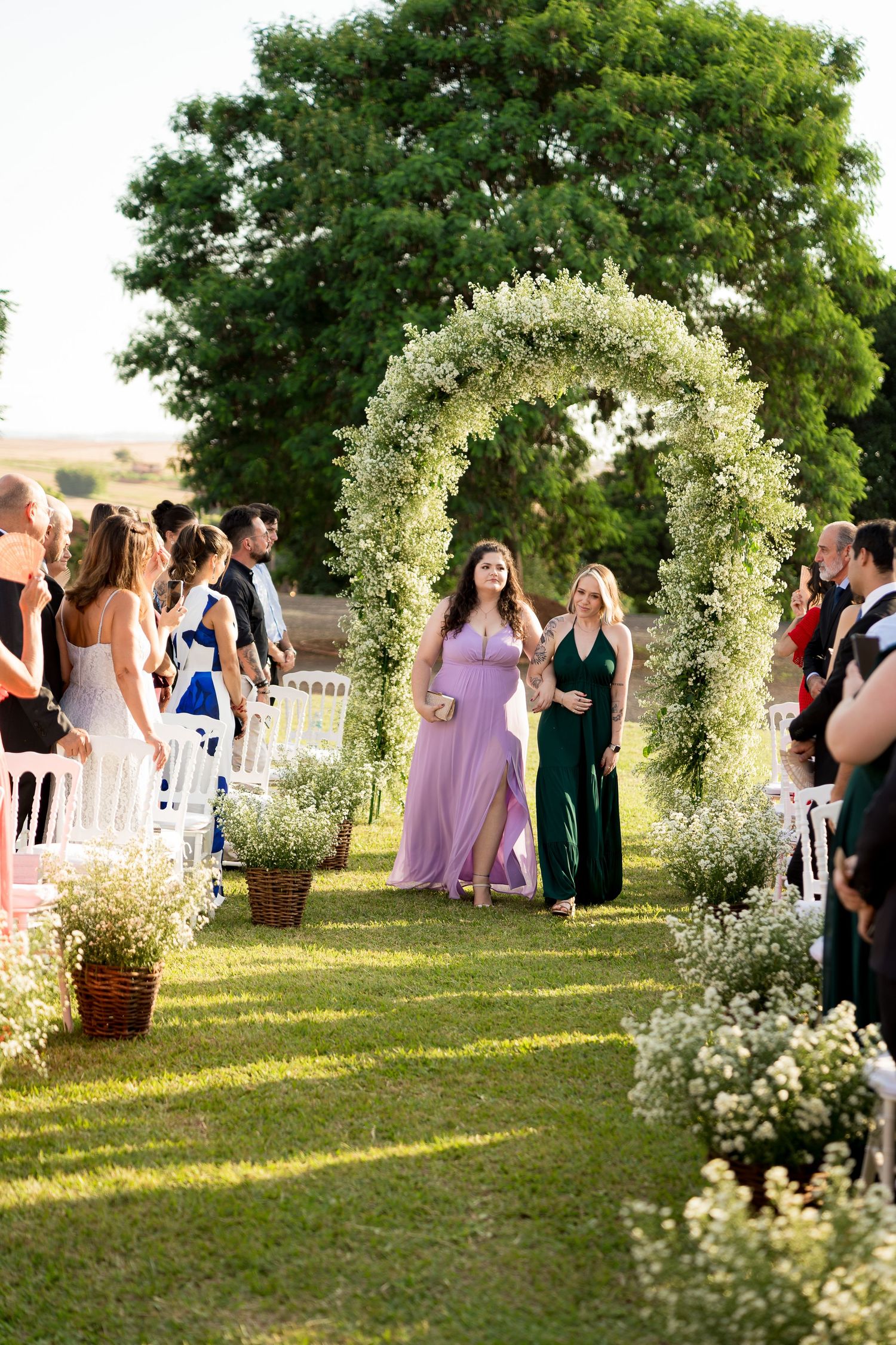 Two bridesmaids walk down a floral arch-adorned outdoor wedding aisle, guests seated on white chairs beside them.