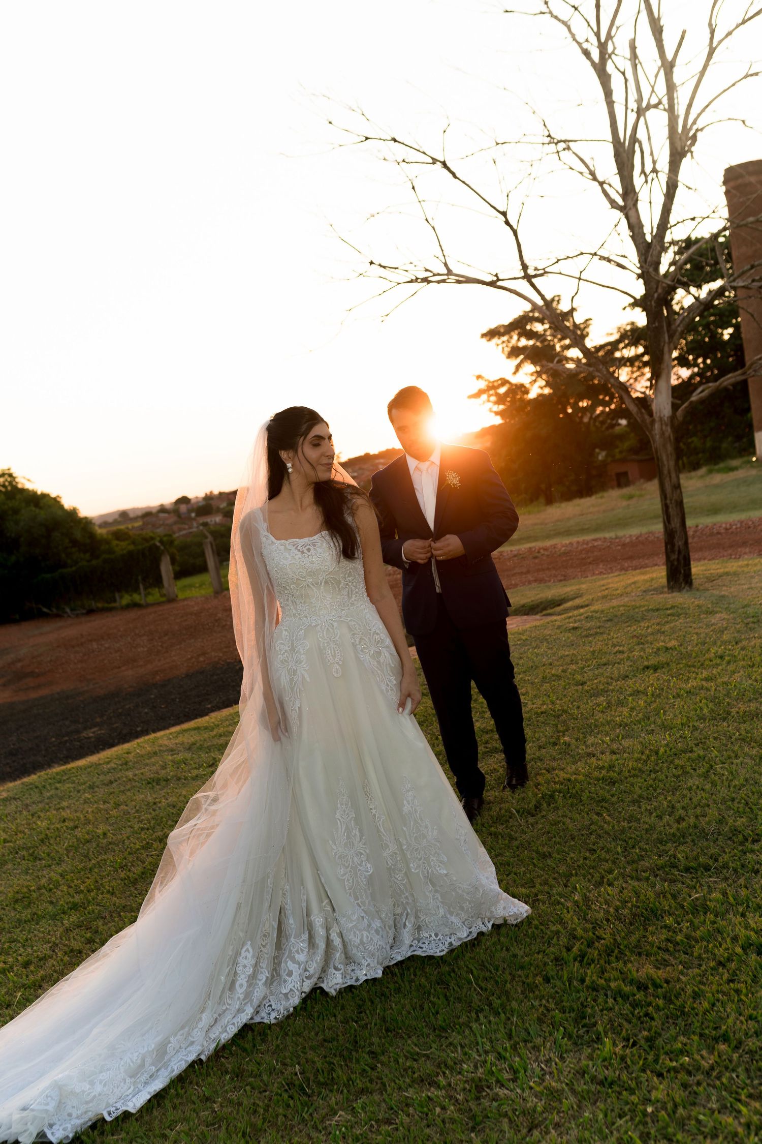 Bride in lace gown and groom in black suit walk at sunset on green lawn near bare tree at golden hour.