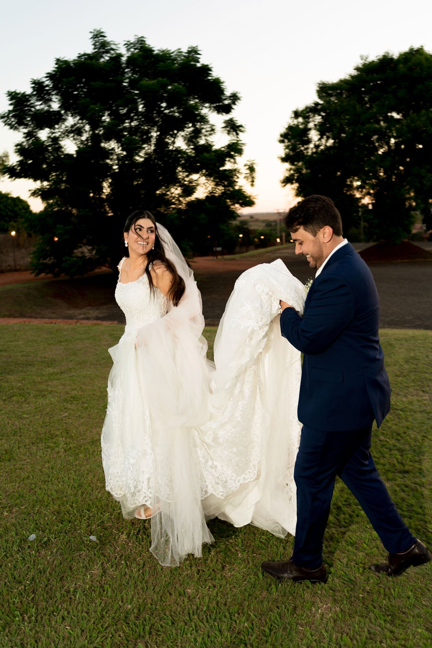 Bride in white lace gown with veil walks outdoors at dusk as groom in navy suit carries her train.