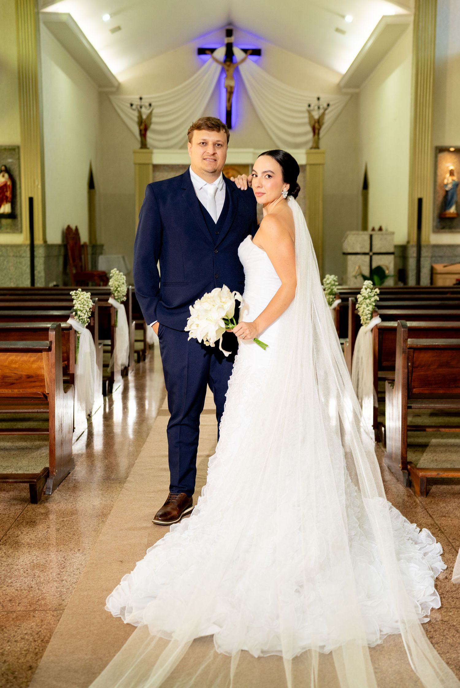 Bride and groom pose together in a church aisle, she in a white gown with veil, he in a navy suit.