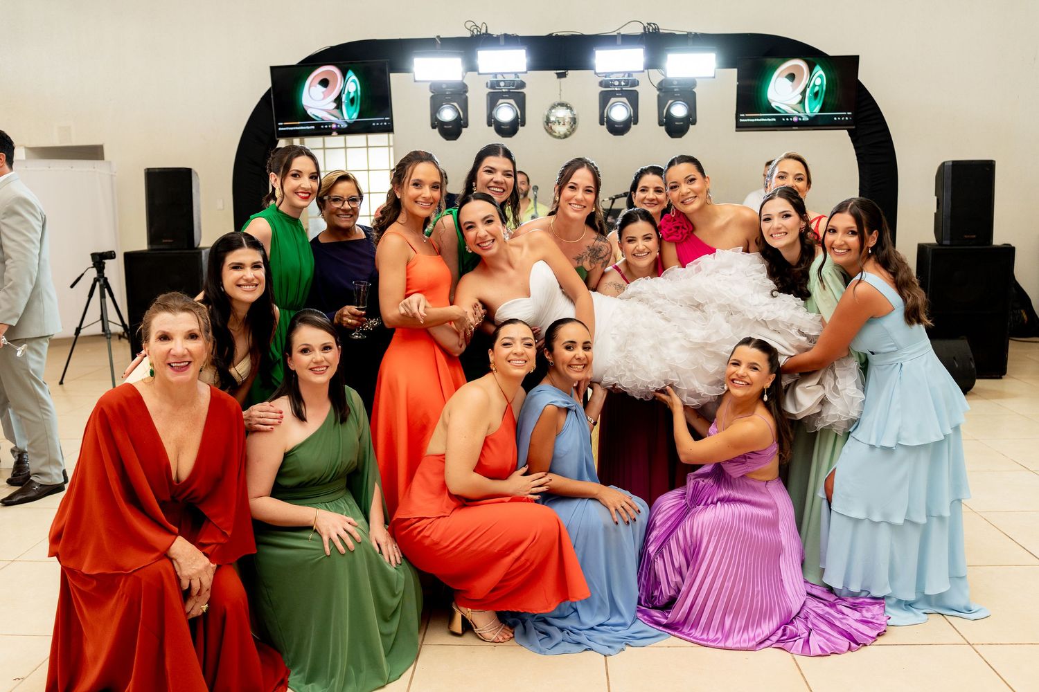 Bride surrounded by bridesmaids in colorful dresses posing together at a wedding reception dance floor.