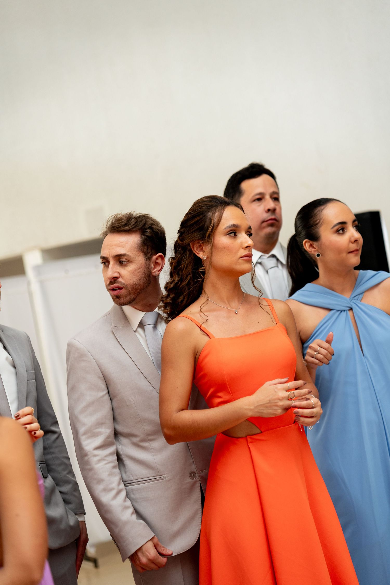 Elegantly dressed guests in orange and blue gowns attend a formal event, standing attentively in a bright room.