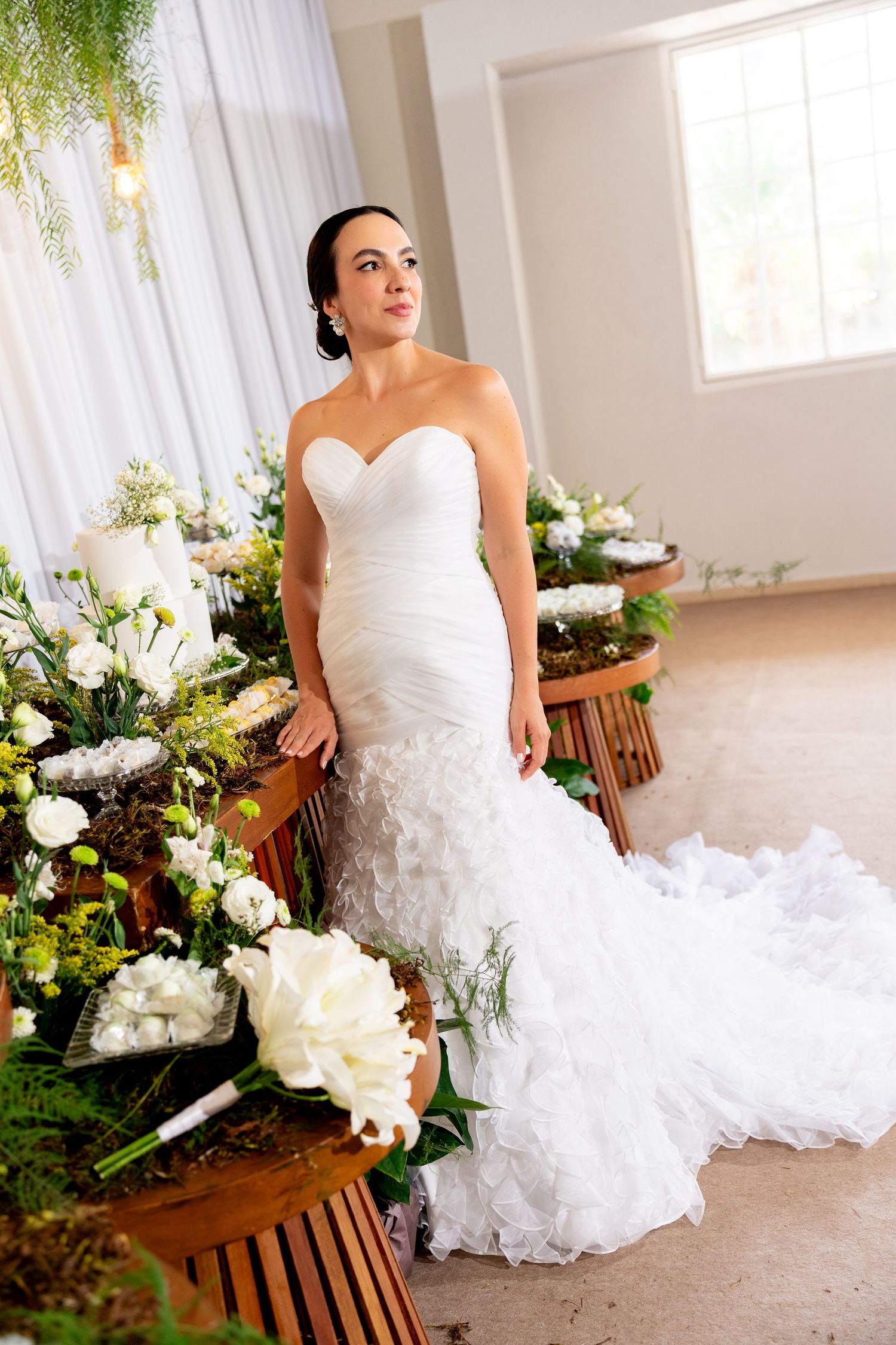 Elegant bride in a strapless mermaid wedding gown poses beside lush floral arrangements with white blooms indoors.
