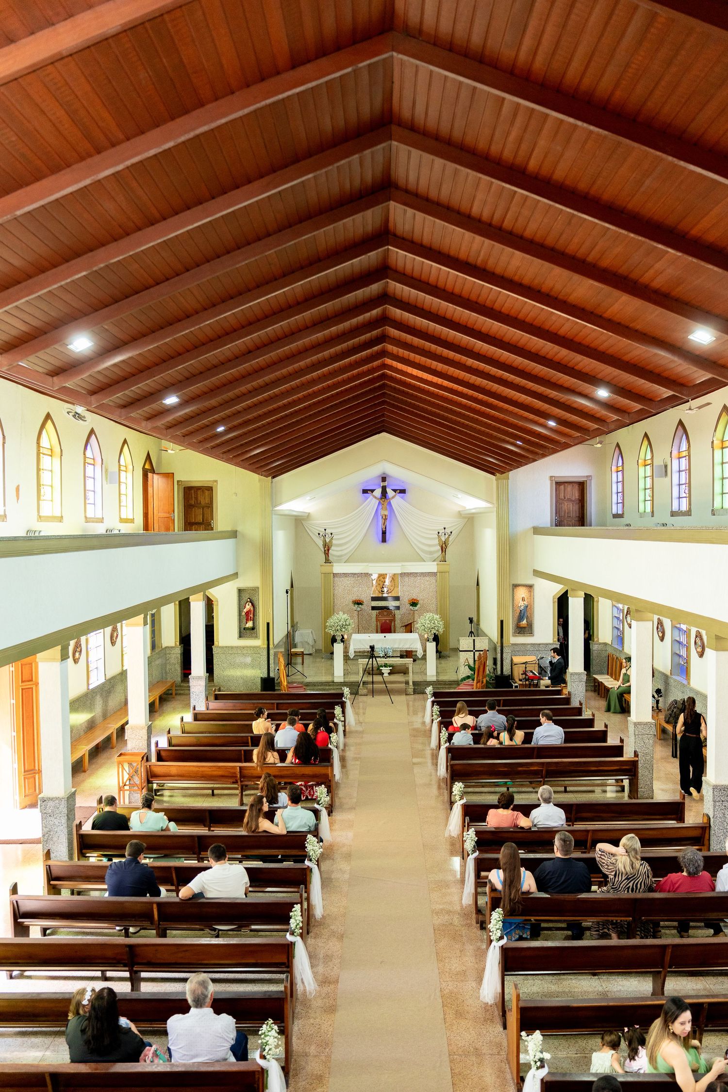 Interior view of a church with wooden ceiling, pews filled with worshippers, and a colorful altar at the front.