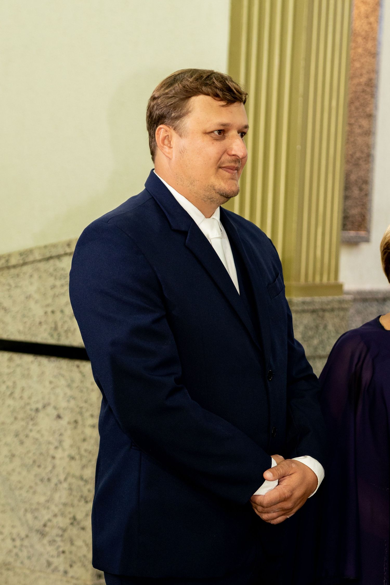 Man in navy blue suit jacket standing indoors near marble columns at a formal event.