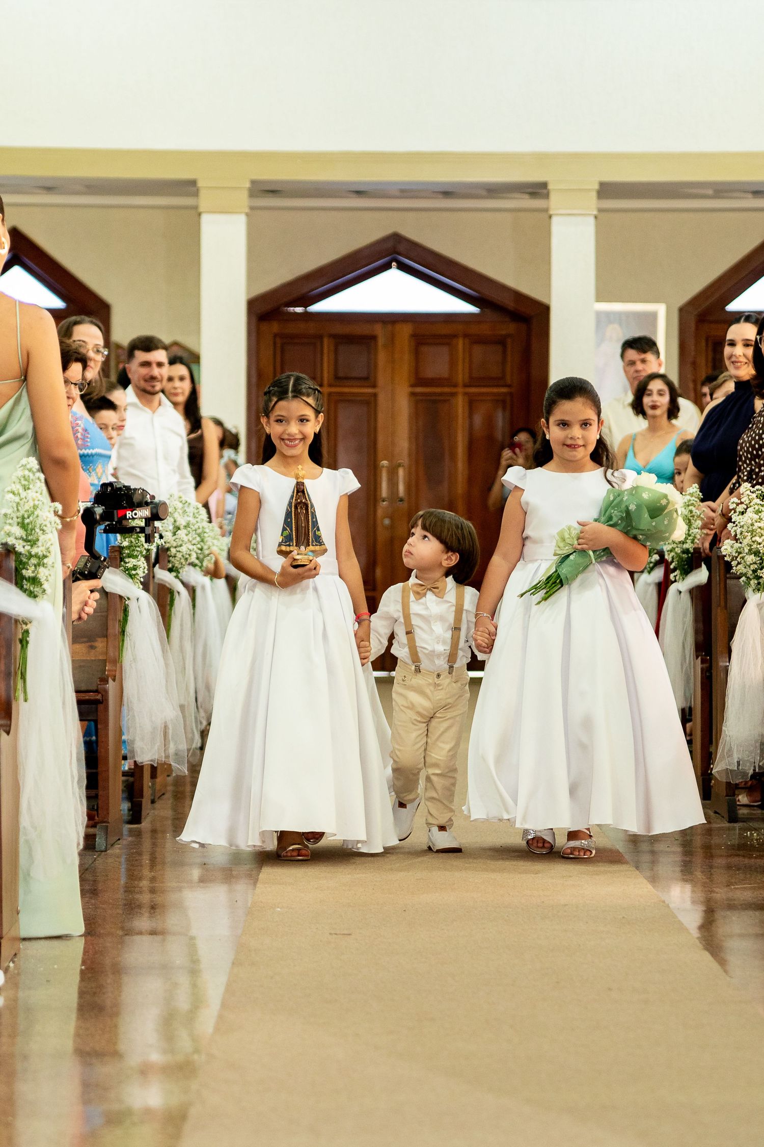 Two flower girls in white dresses and a ring bearer walk down a church aisle during a wedding ceremony.