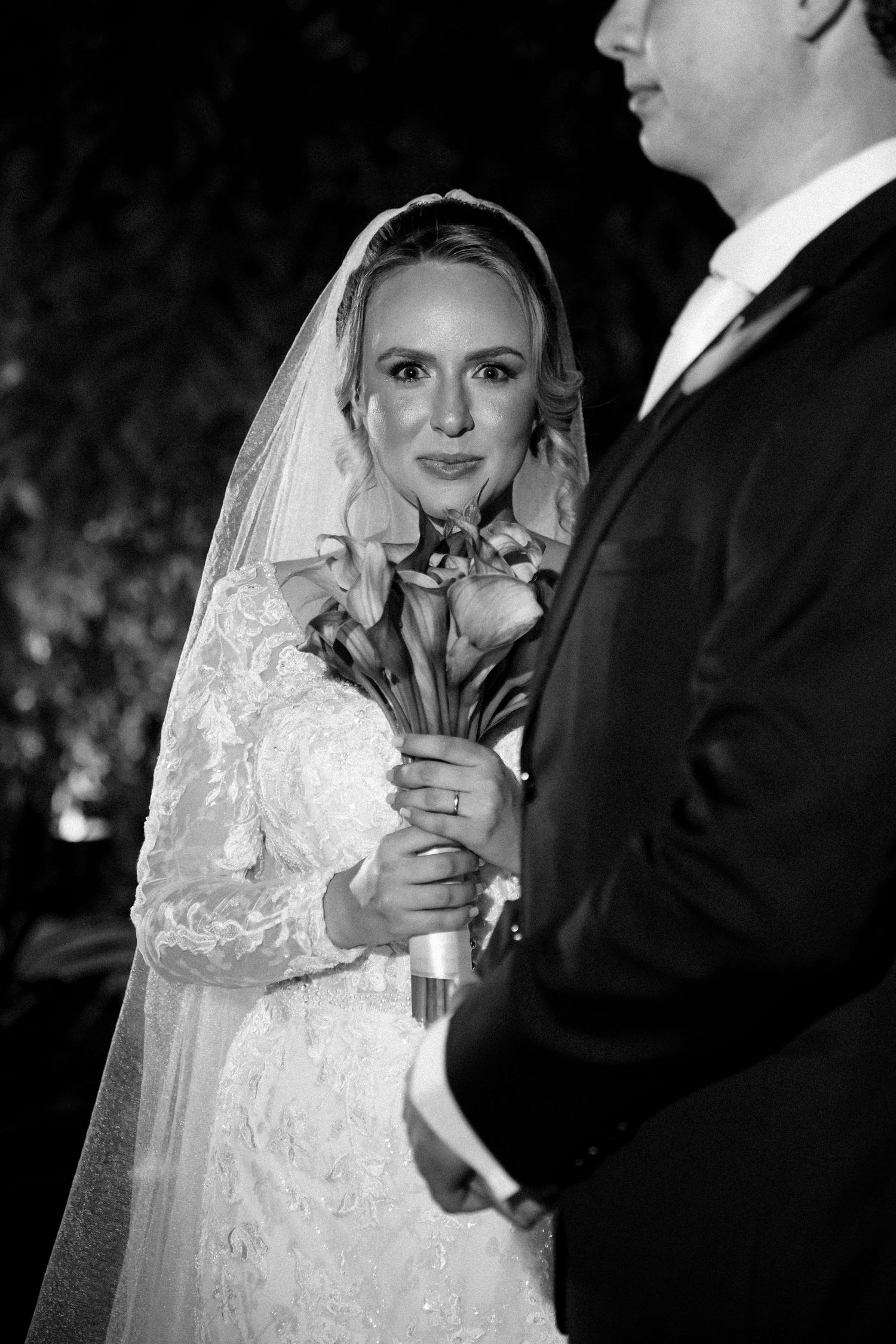 Black and white wedding photo of a bride in lace gown holding bouquet, gazing at groom during ceremony.