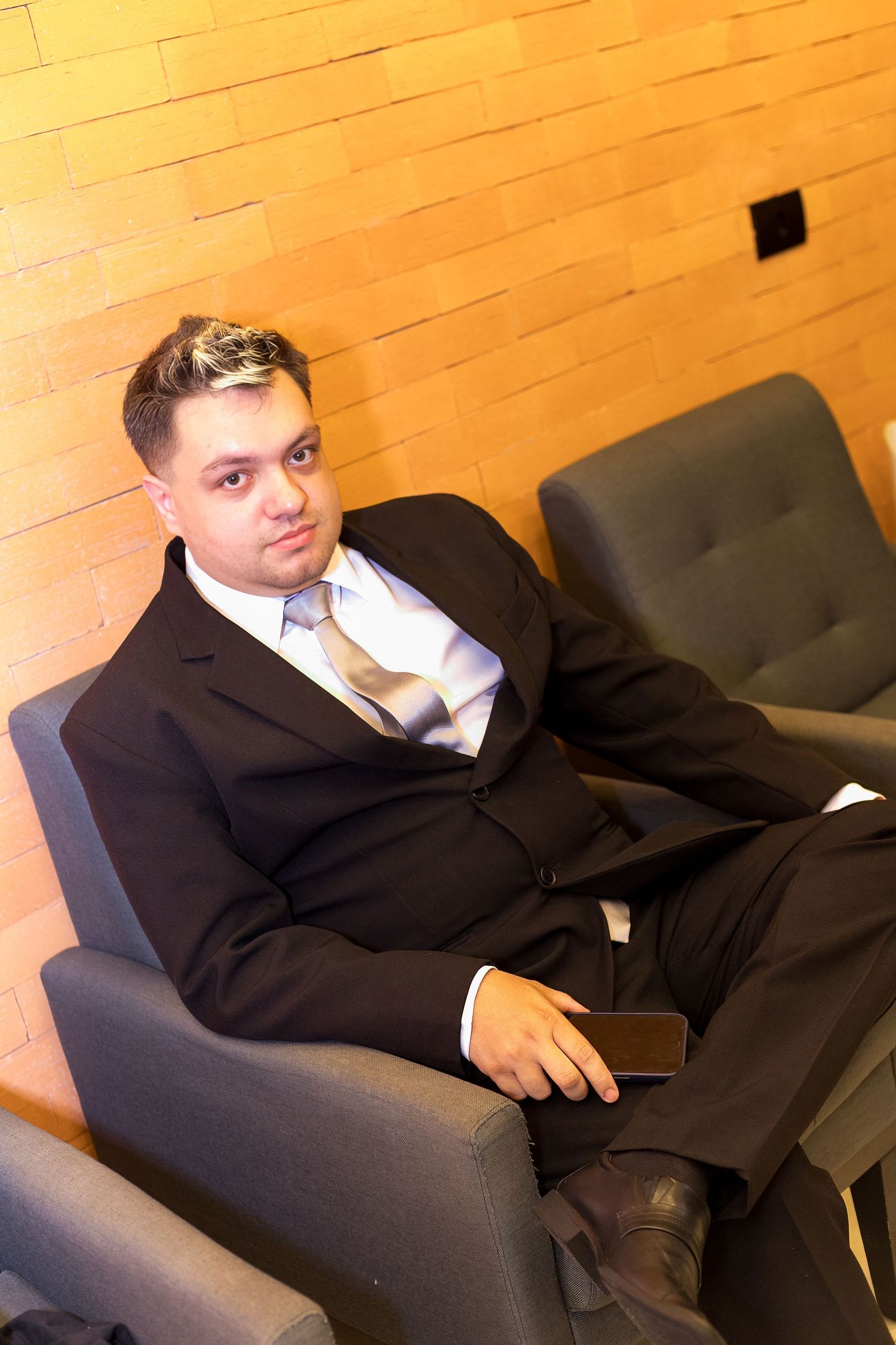 A man in a dark suit and gold tie sits relaxed in a gray armchair, holding a phone, against a warm brick wall.