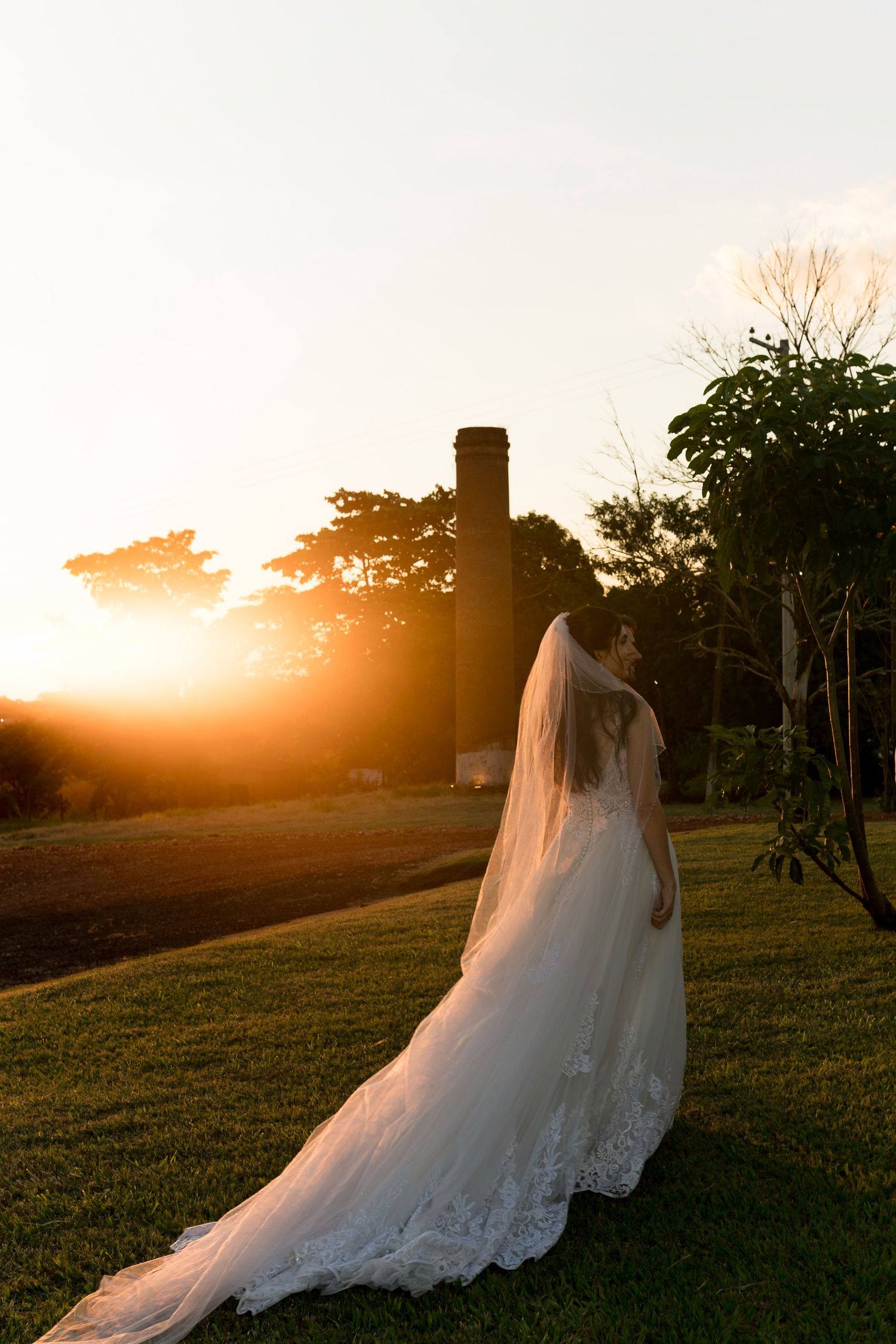 Bride in white lace gown and veil stands on green lawn at golden sunset with chimney silhouette backdrop.