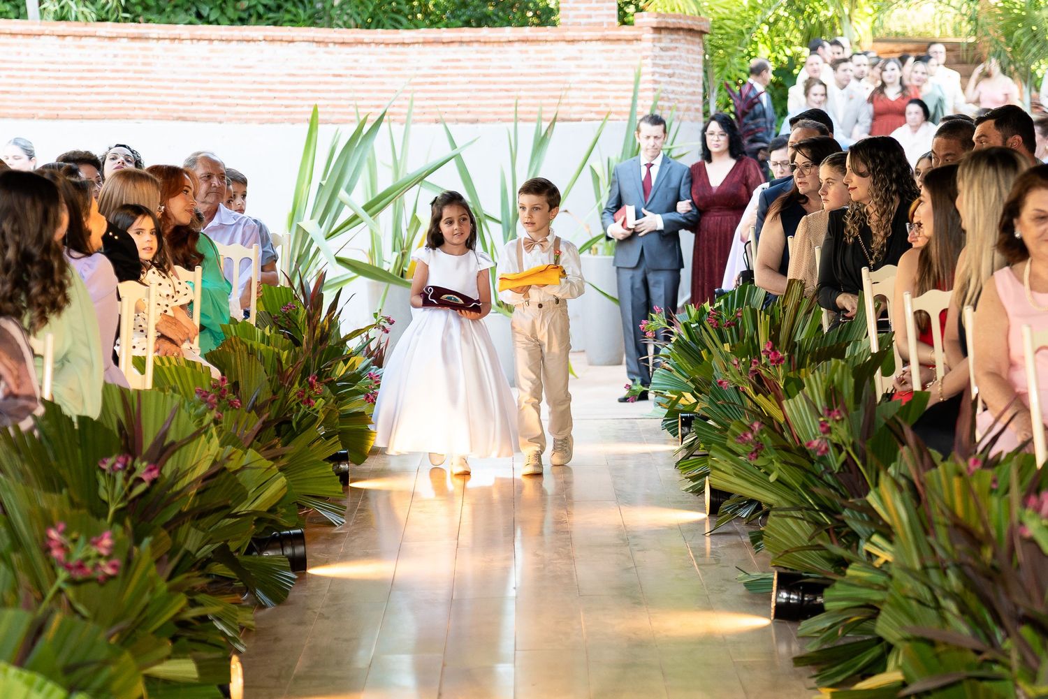 Flower girl and ring bearer walking down wedding aisle lined with tropical plants during outdoor ceremony.