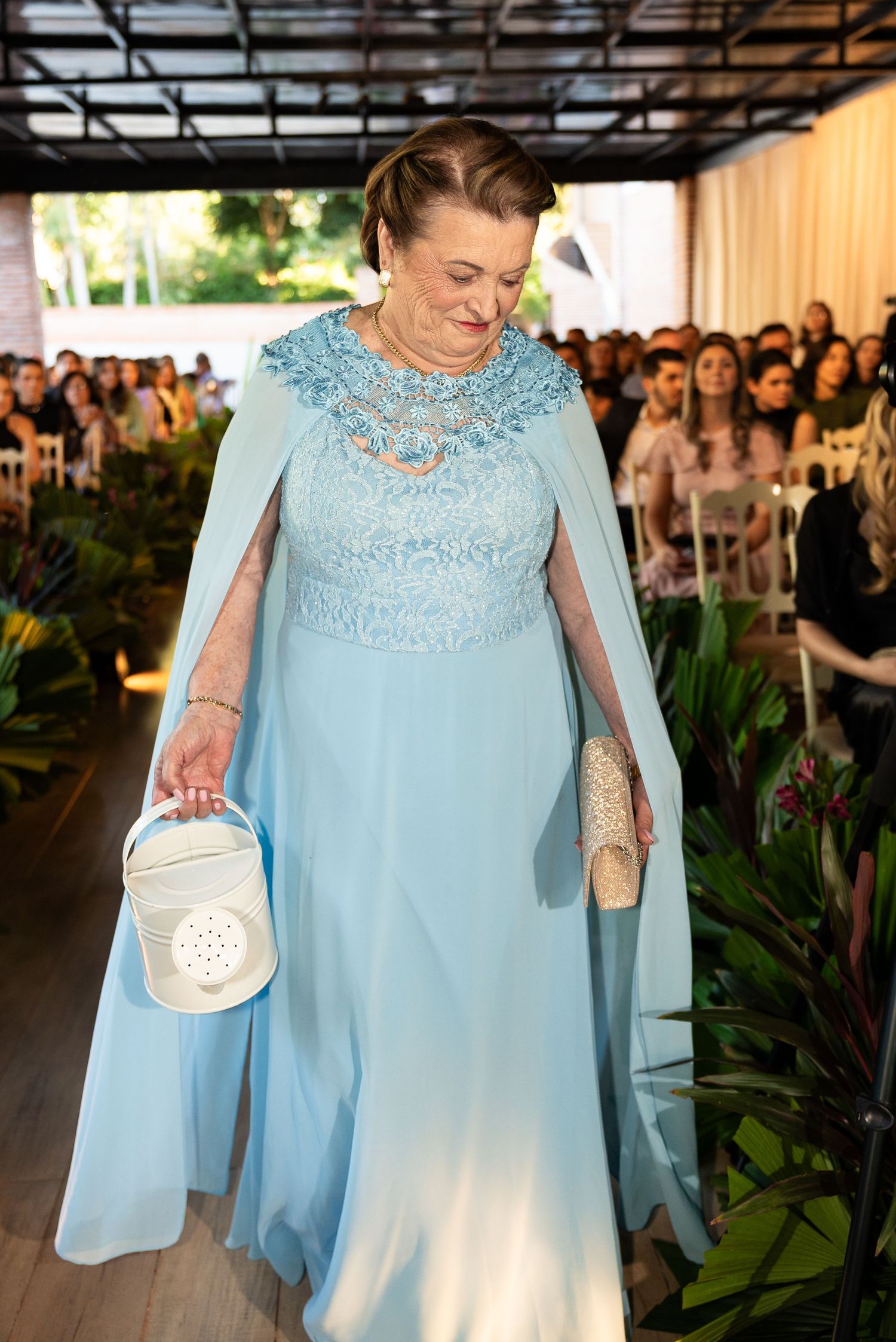 Elderly woman in elegant light blue lace cape gown walking down wedding aisle carrying white handbag.