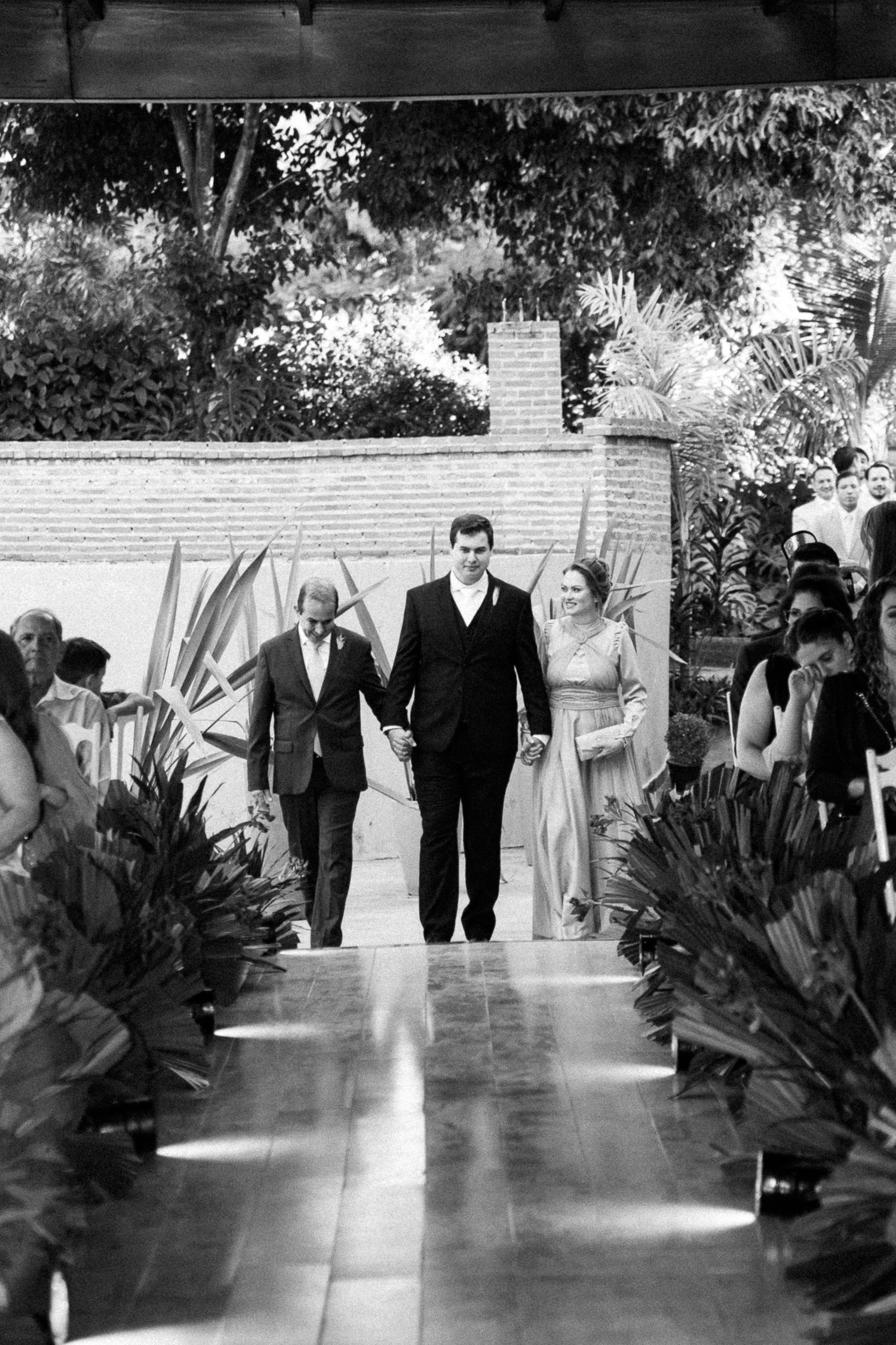 Groom walks down wedding aisle escorted by parents in black and white outdoor ceremony photo.