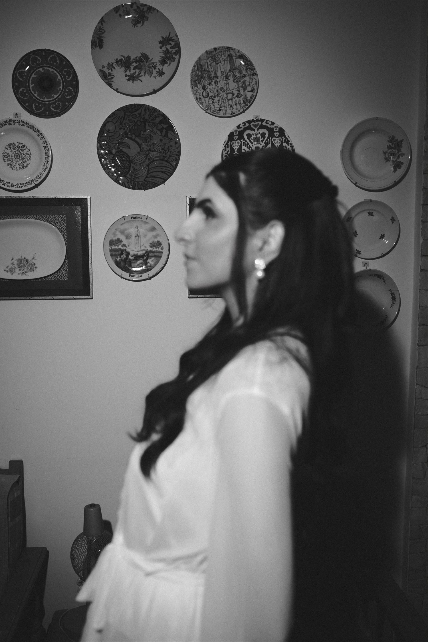Black and white photo of a bride in white dress with long dark hair, standing before a wall of decorative plates.