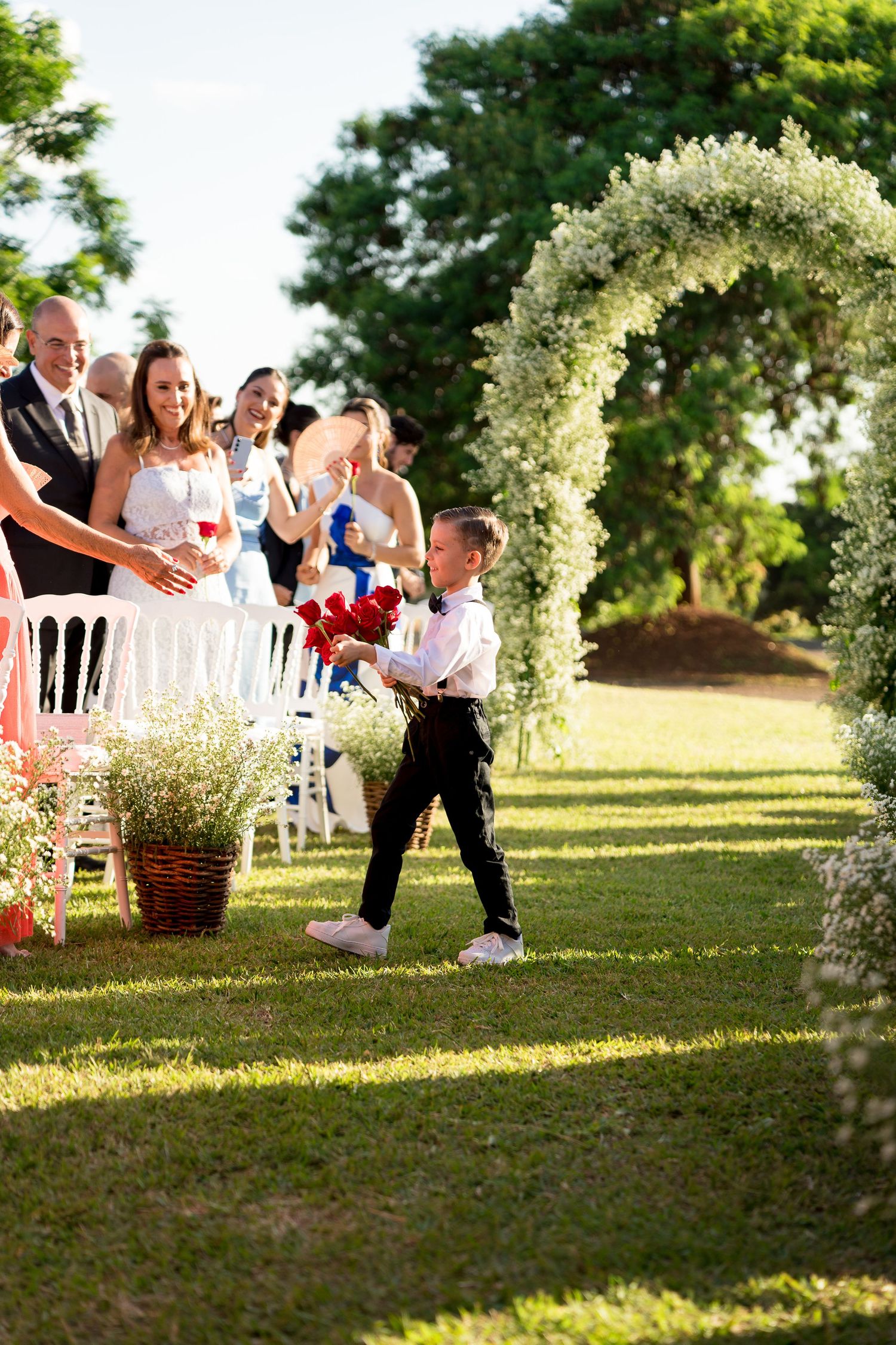 Young ring bearer walks down outdoor wedding aisle holding red flowers, guests applaud beside floral arch.