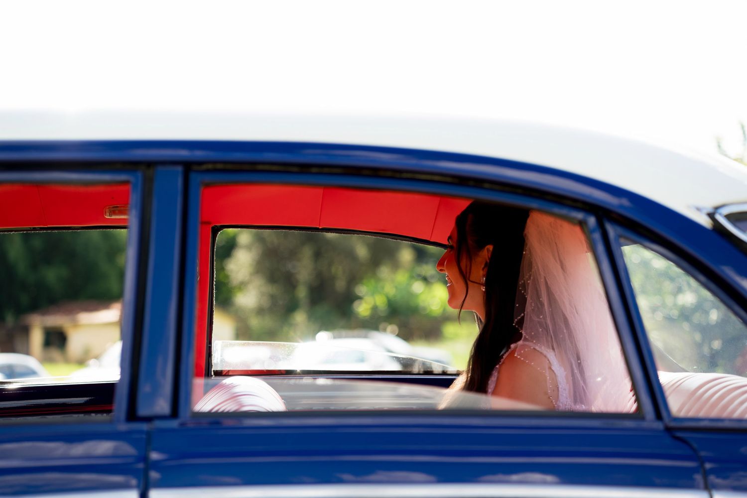 Bride in wedding veil sitting in backseat of vintage blue car, viewed through open window on sunny day.