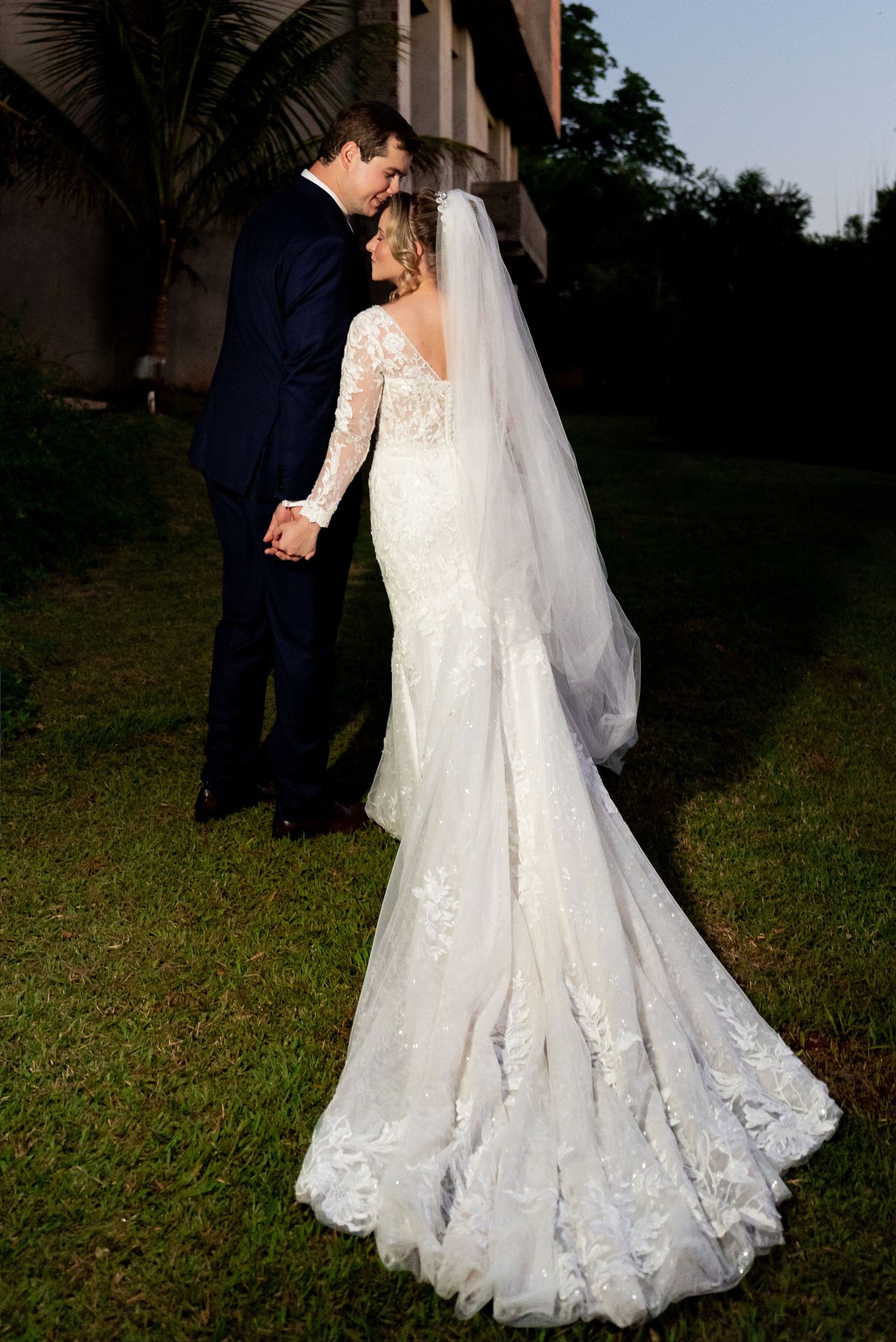 Bride in lace mermaid gown with veil and groom in navy suit hold hands outdoors at golden hour wedding.