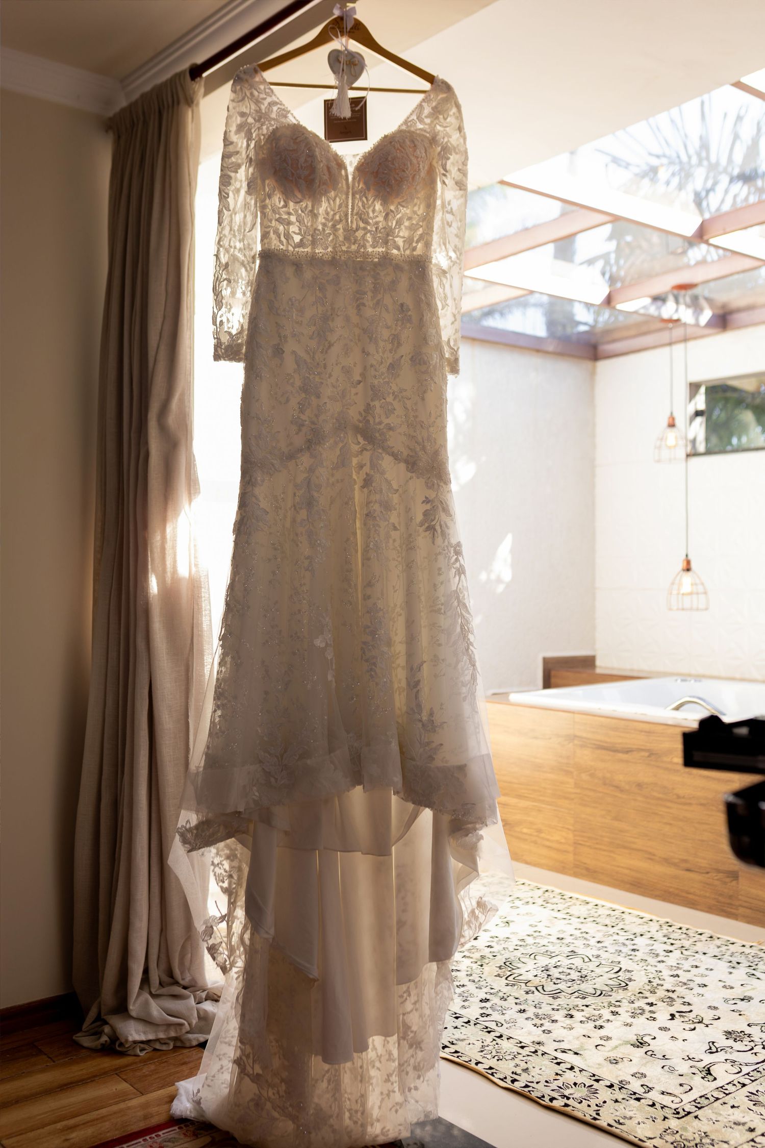 Elegant lace wedding dress with sleeves hanging on a wooden hanger in a sunlit bridal suite with modern decor.