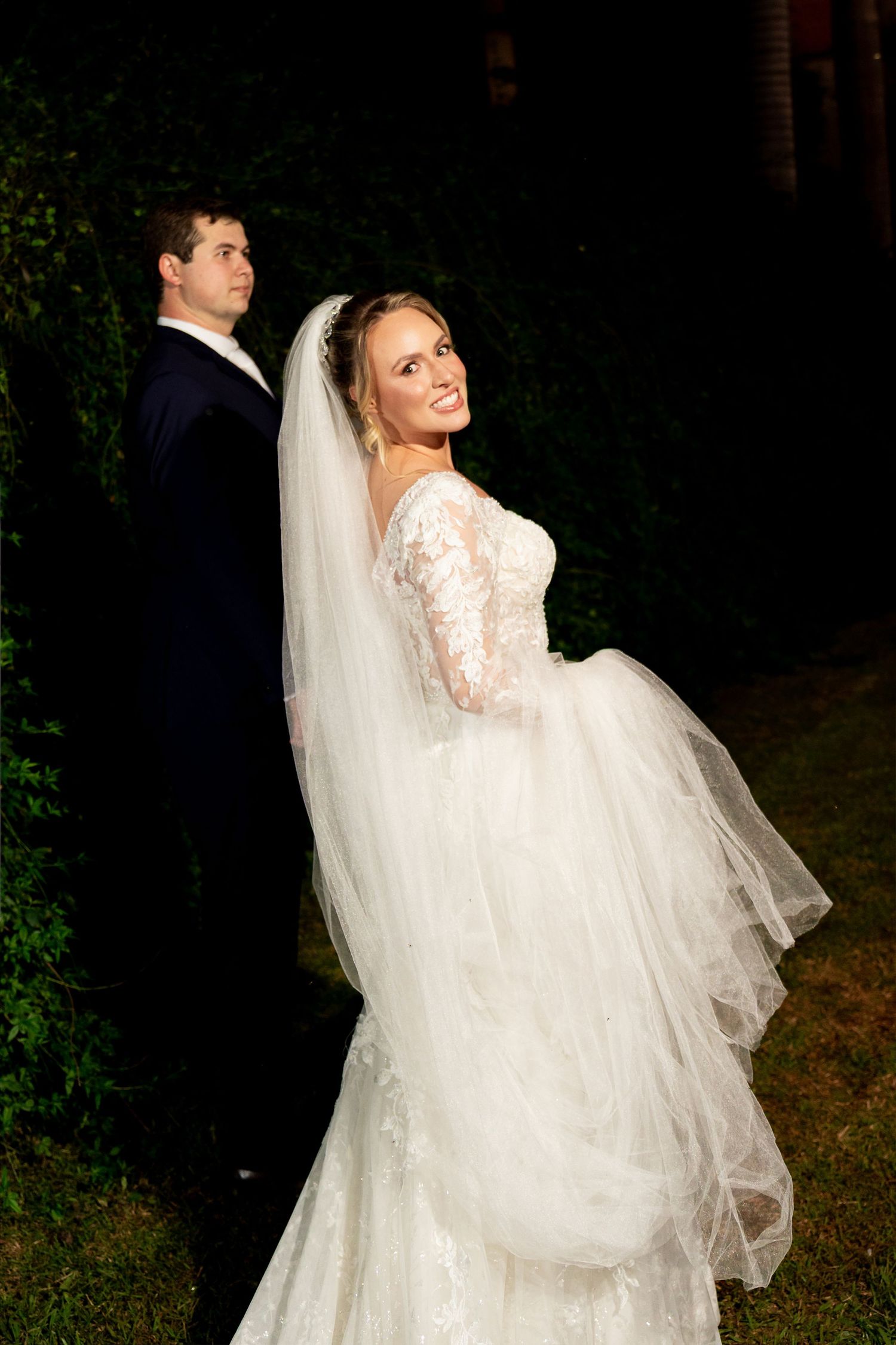 Bride in lace gown and veil smiles over shoulder beside groom in tuxedo during outdoor night wedding portrait.