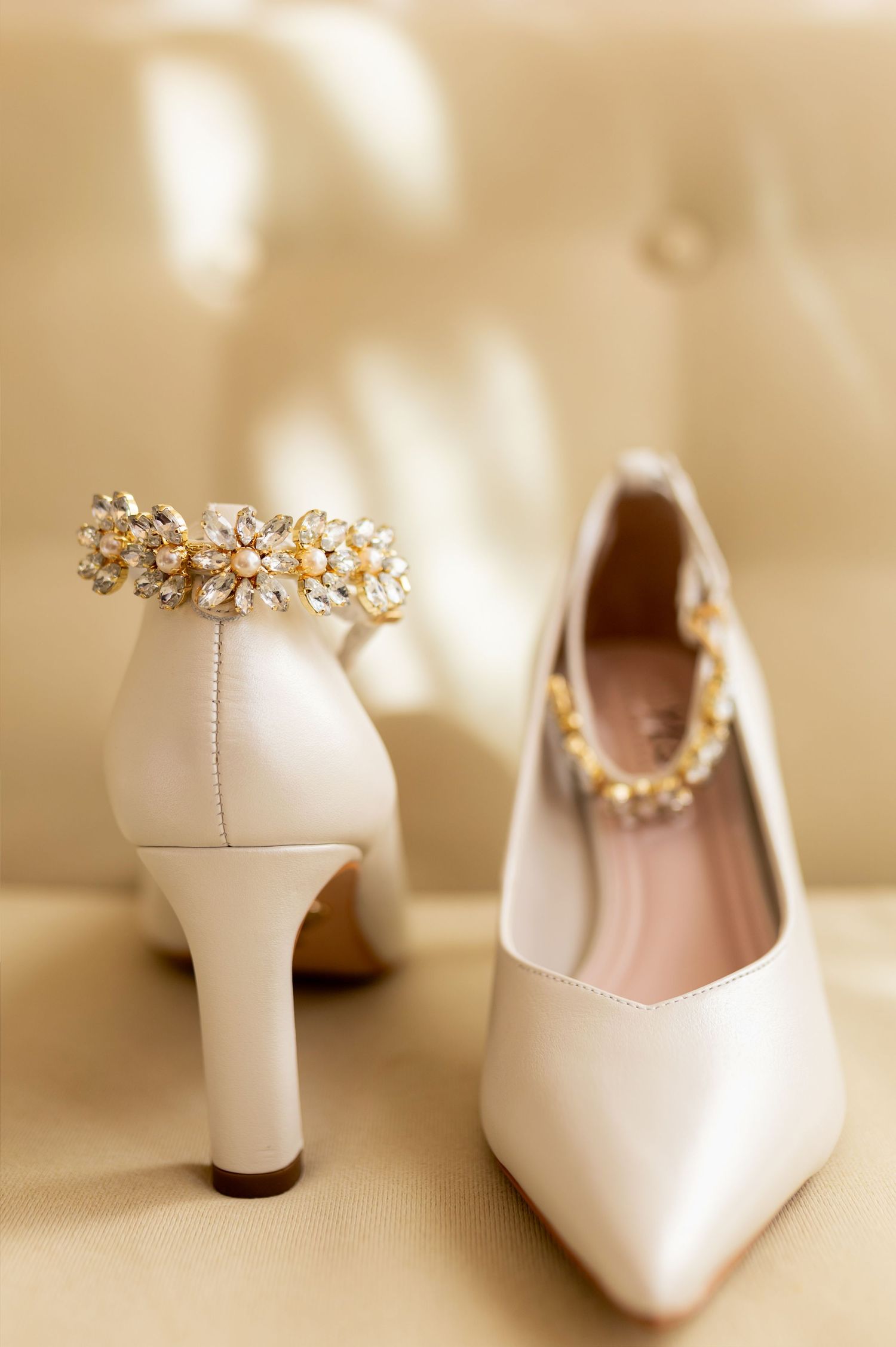 Elegant white bridal heels with gold crystal floral embellishments displayed on a cream leather surface.