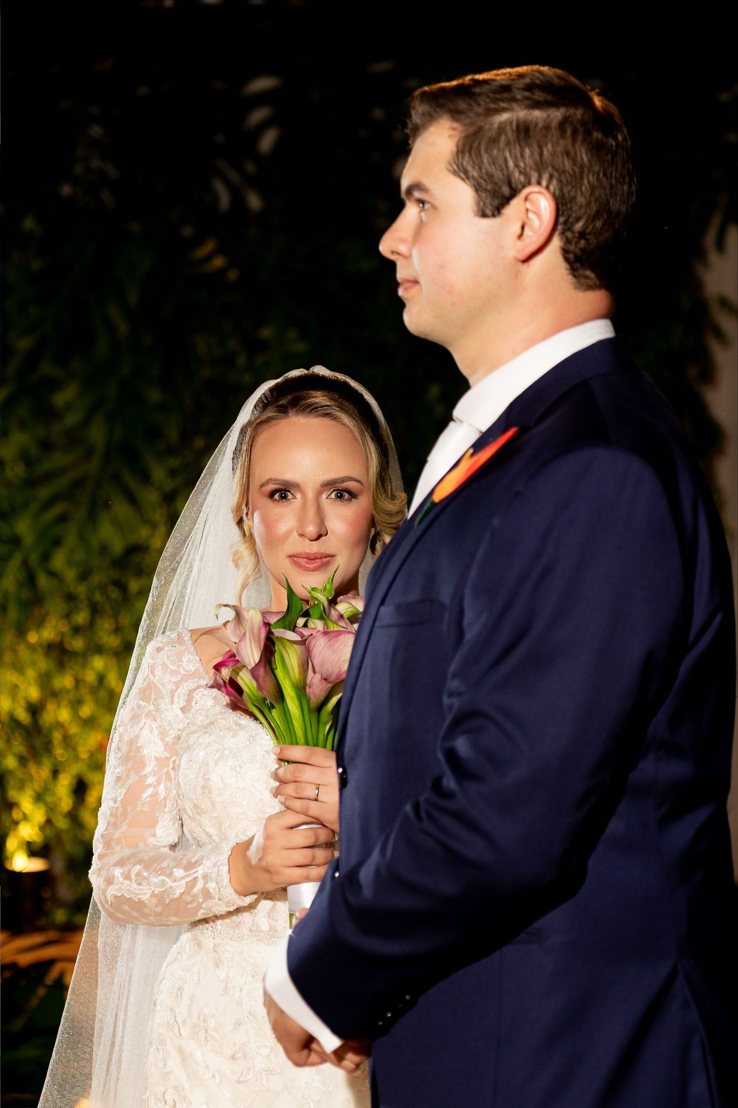 Bride in lace gown holding pink calla lily bouquet stands beside groom in navy suit at outdoor evening wedding ceremony.