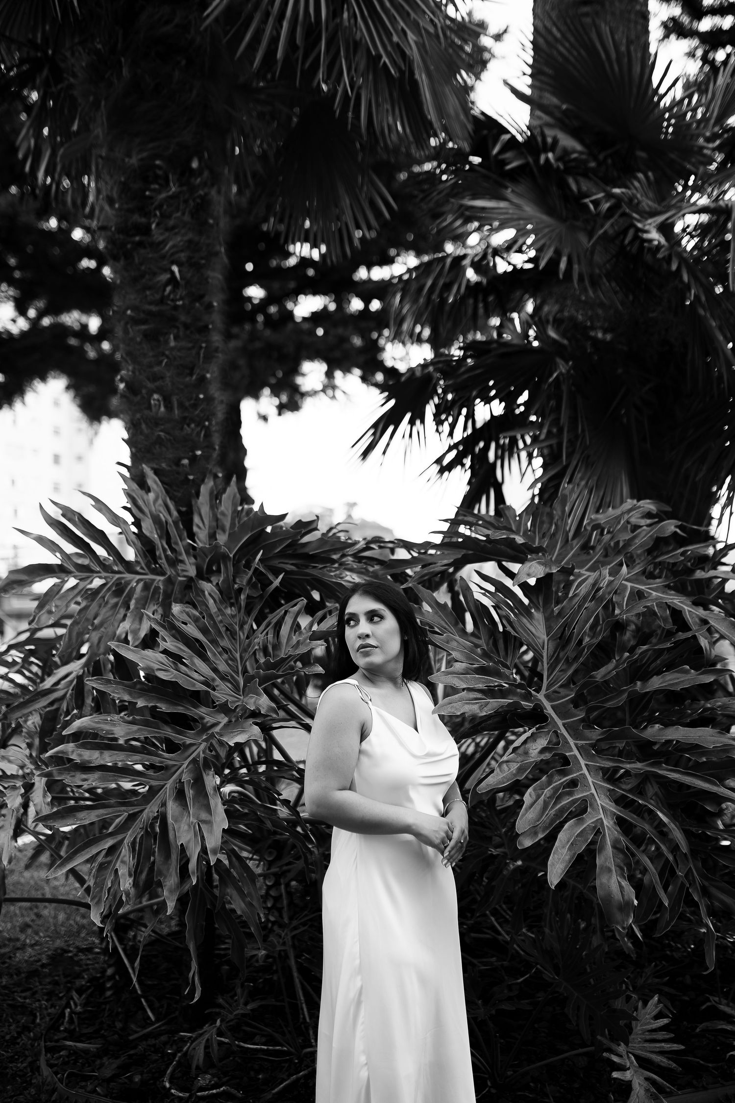 Black and white portrait of woman in white dress posed before tropical palm trees and large leaves.