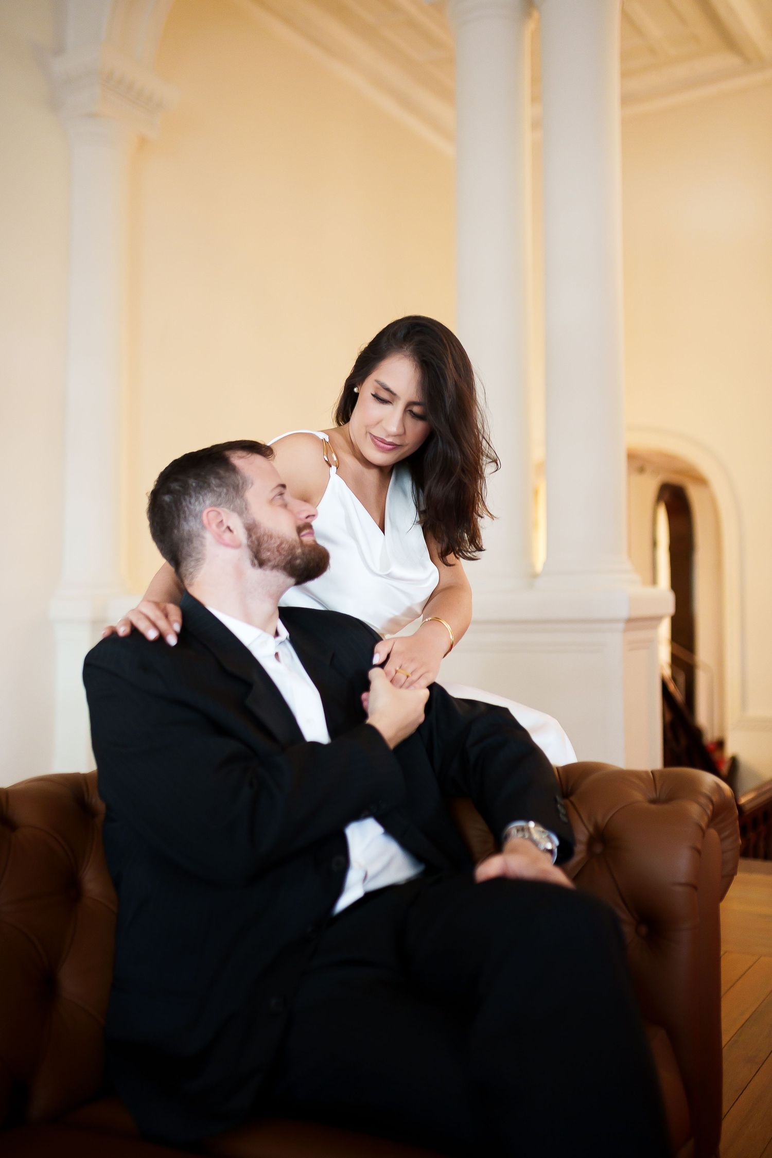 Elegant couple sharing a romantic moment on a leather sofa in a classic interior, man in black suit, woman in white dress.