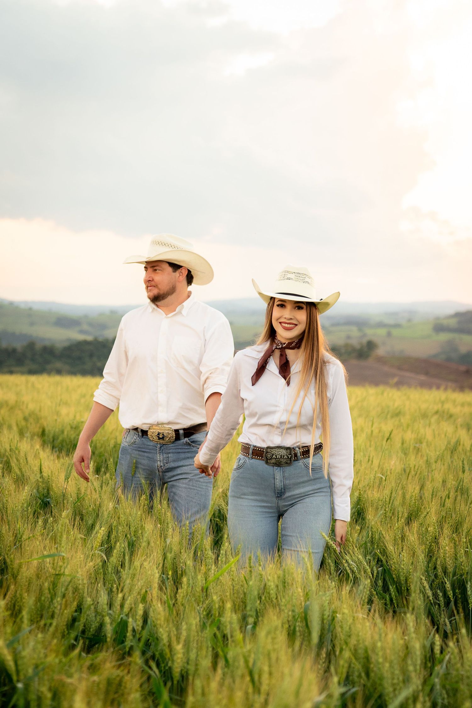 Couple in white shirts and cowboy hats walking through a green wheat field at sunset.