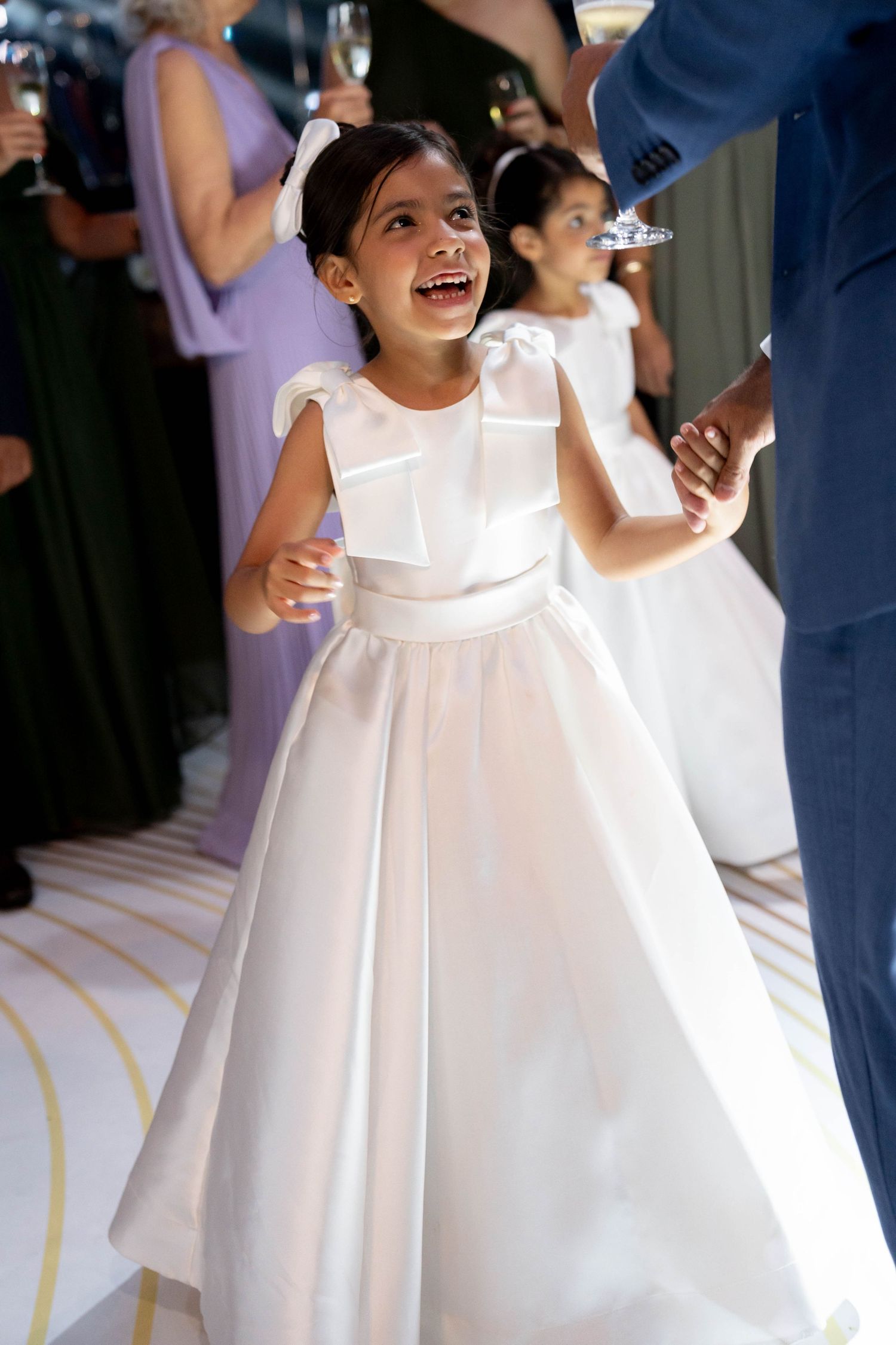 Joyful flower girl in white ball gown dress laughing and holding hands at an elegant wedding reception.