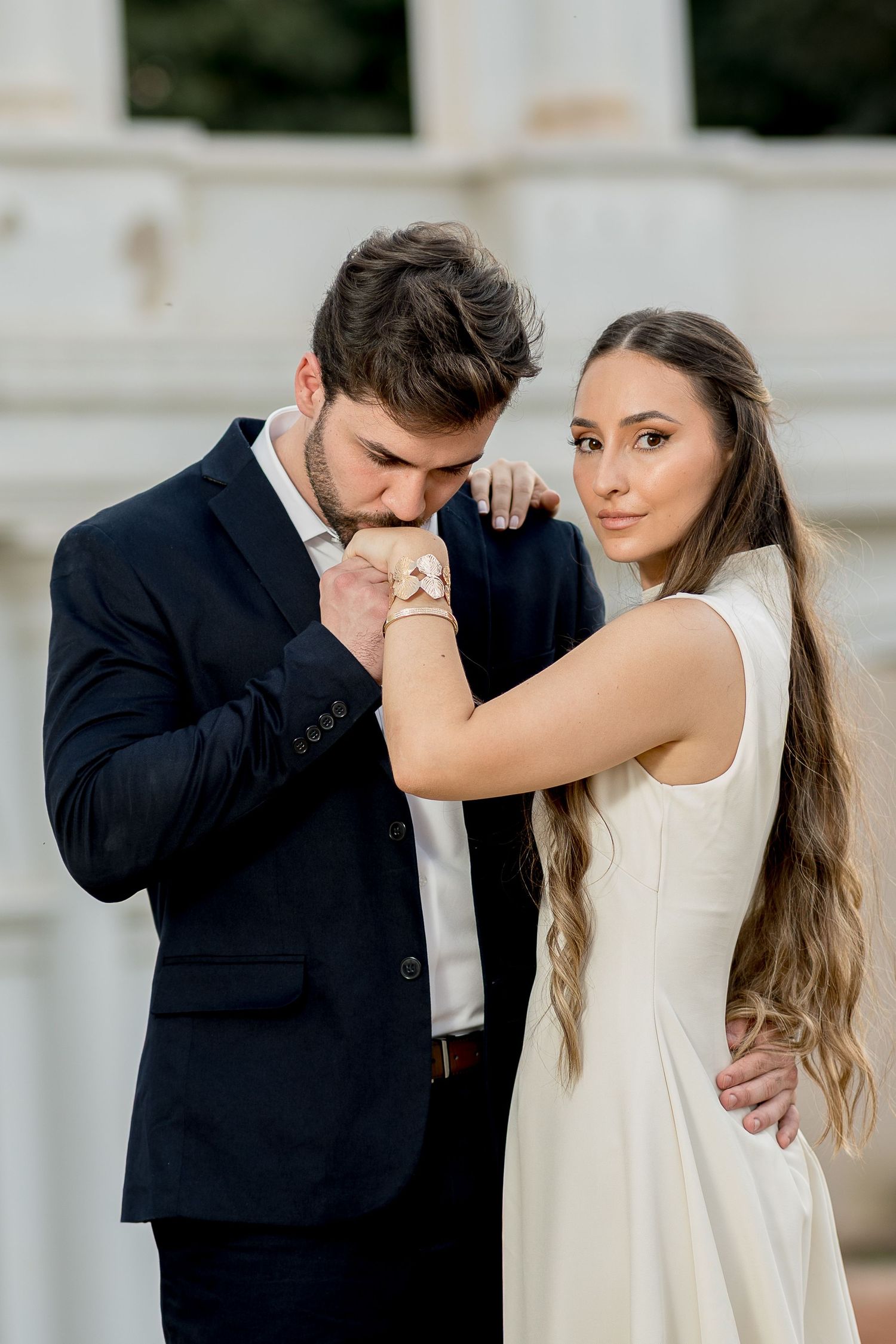 A groom in a dark suit kisses his bride's hand as she gazes forward in a white sleeveless dress.