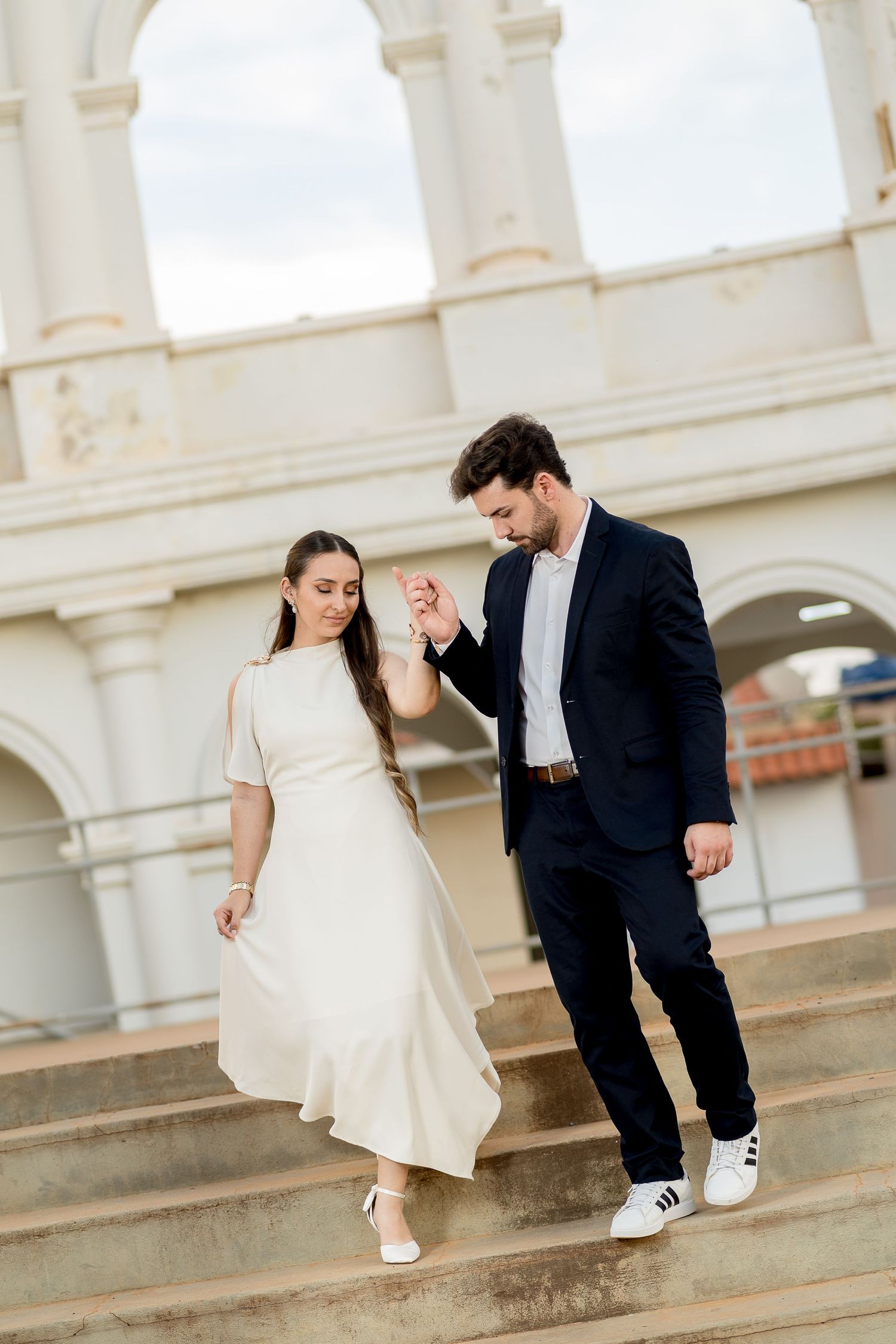 Elegant couple descending outdoor steps, woman in white dress, man in navy suit holding hands at a classical building.