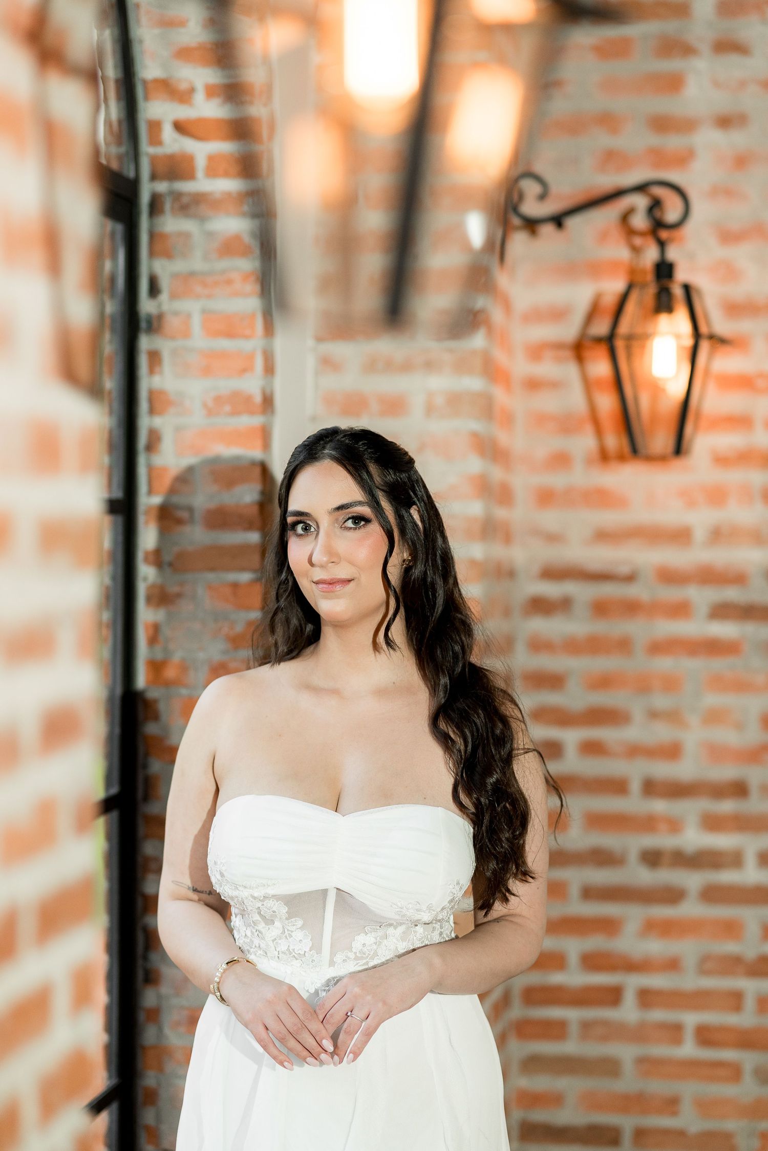 Bride in strapless white corset gown with long wavy hair poses near brick wall with vintage lantern.