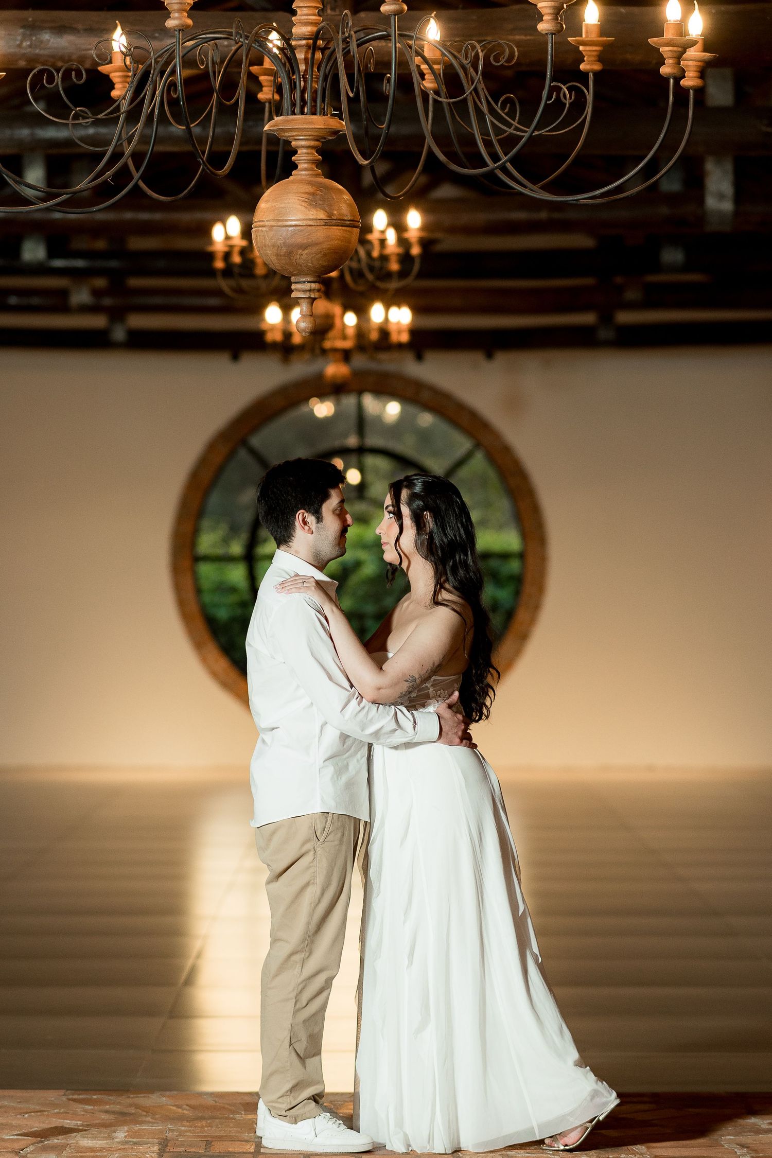 Bride and groom share romantic first dance under elegant chandelier in front of circular window at wedding venue.